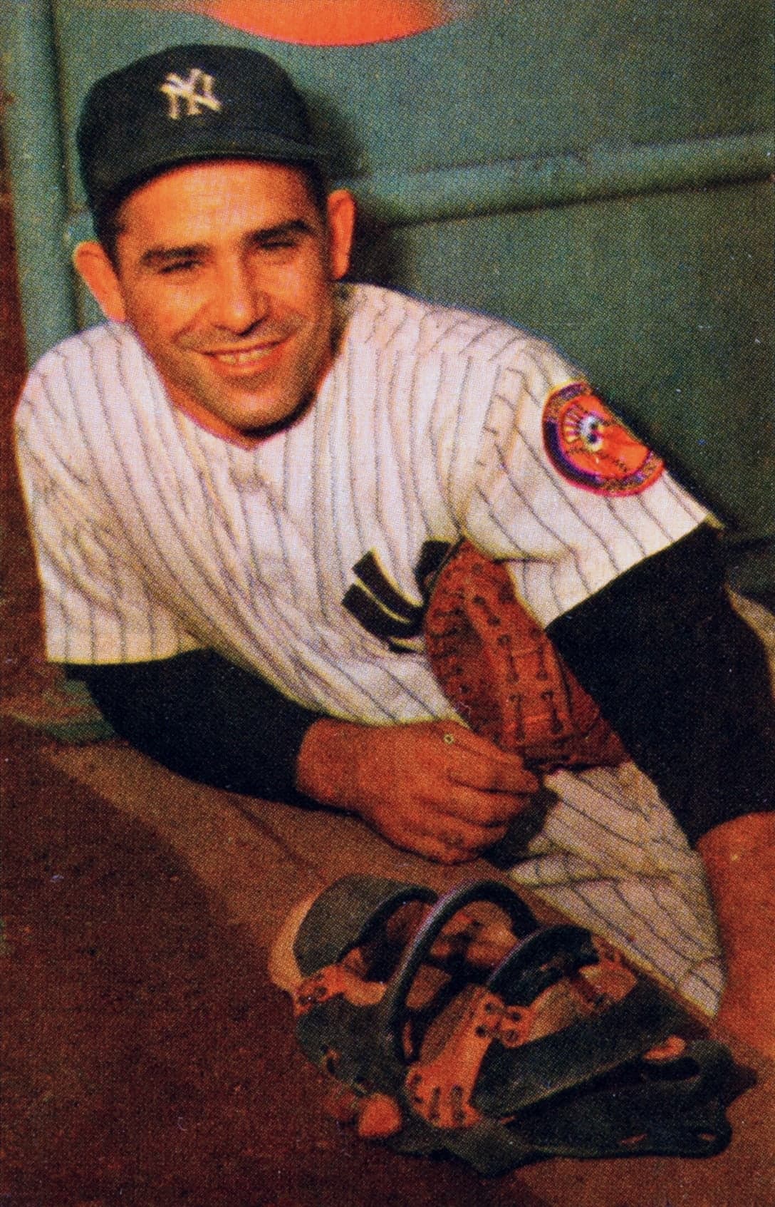 Black-and-white photograph of a baseball player in a dark cap and light uniform, crouching in catcher's gear, one hand resting on his knee, the other holding a catcher's mitt, with stadium seating visible in the background.