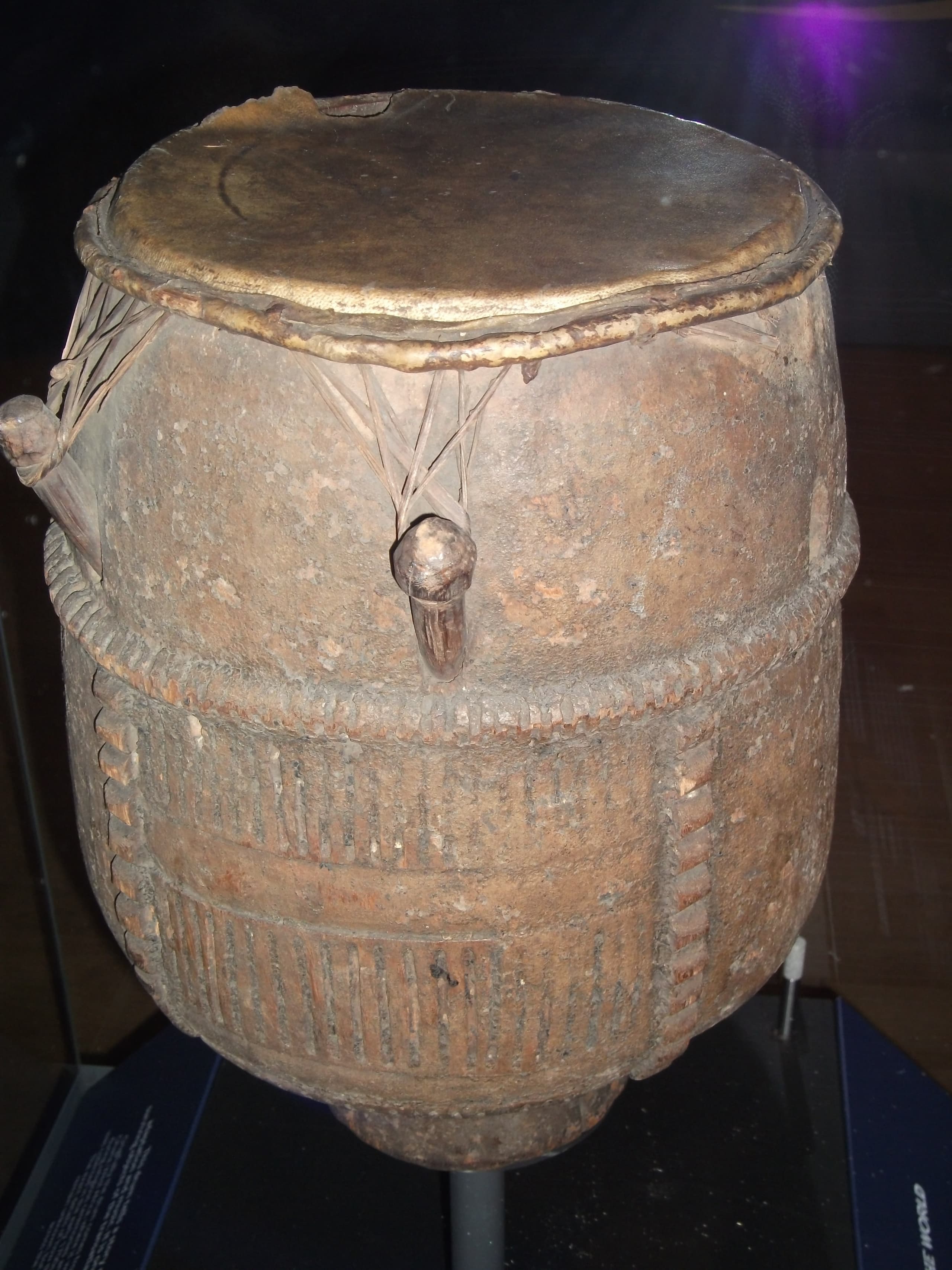 A wooden drum, carved from a single piece of timber, with a skin stretched across the top and held taut by wooden pegs. The surface is worn smooth.