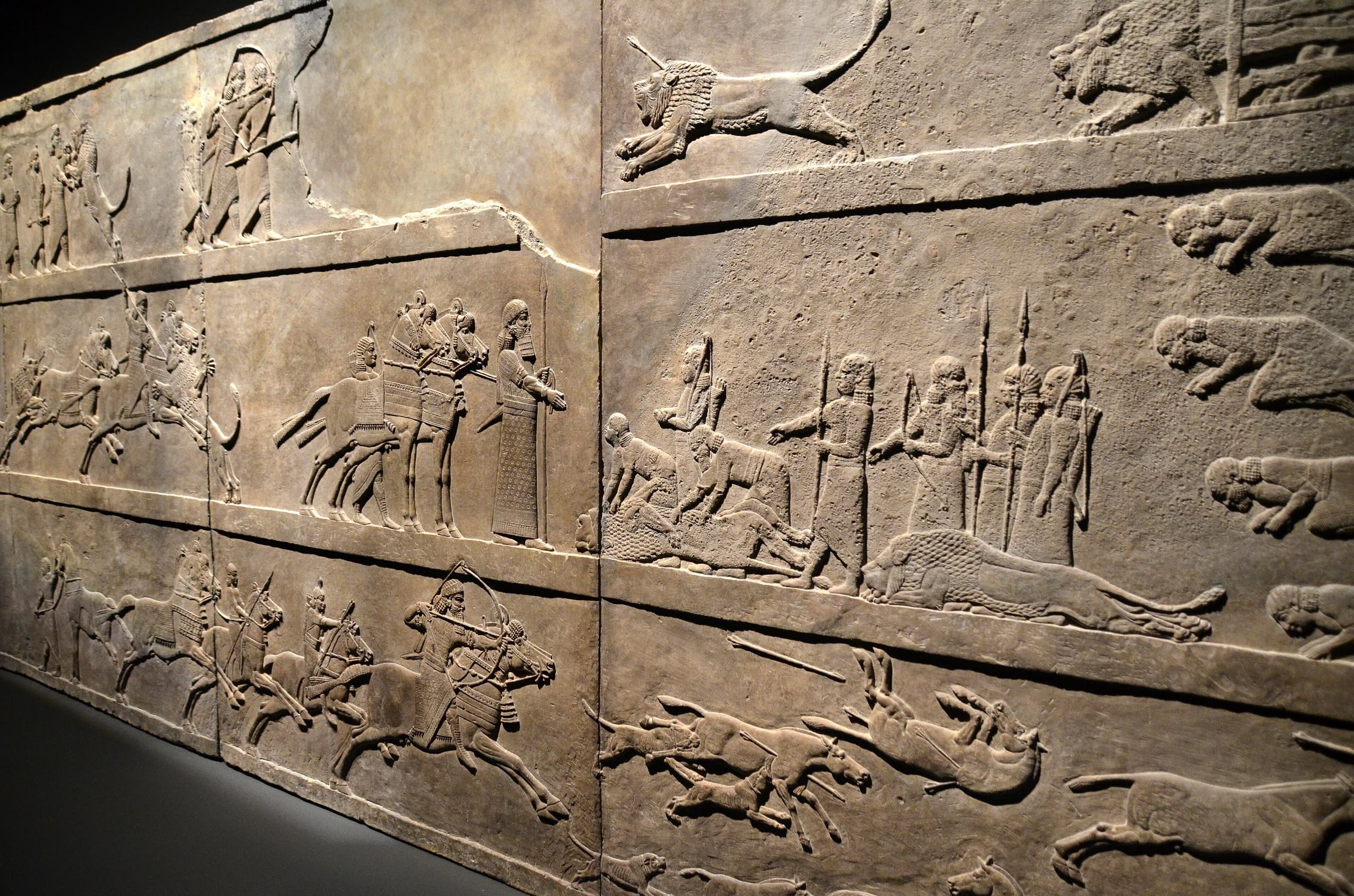 Stone relief panels showing a king hunting lions in an enclosed arena. Lions leap, fall, and die; some are pierced with arrows. One lioness drags her paralysed hind legs. Attendants and dogs surround the scene.