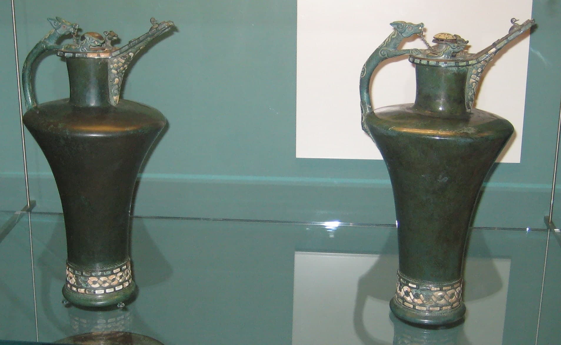 Two bronze wine jugs with swelling bodies, high curved handles, and trefoil spouts, their surfaces covered in geometric patterns, stylised animals, and red coral studs. A small bronze duck sits at the base of each handle.