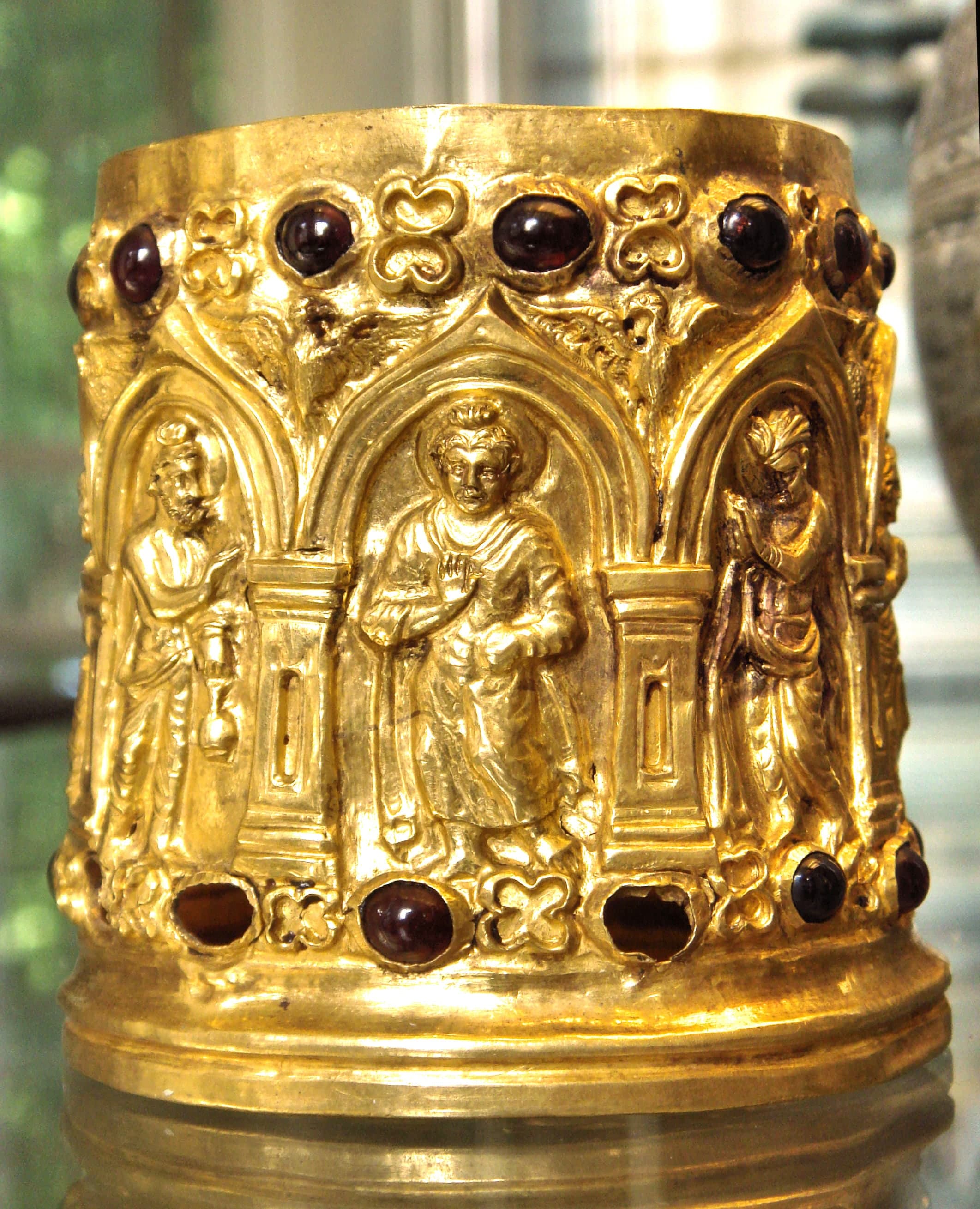 A small cylindrical gold reliquary with repoussé decoration showing standing figures in draped robes within arched niches.