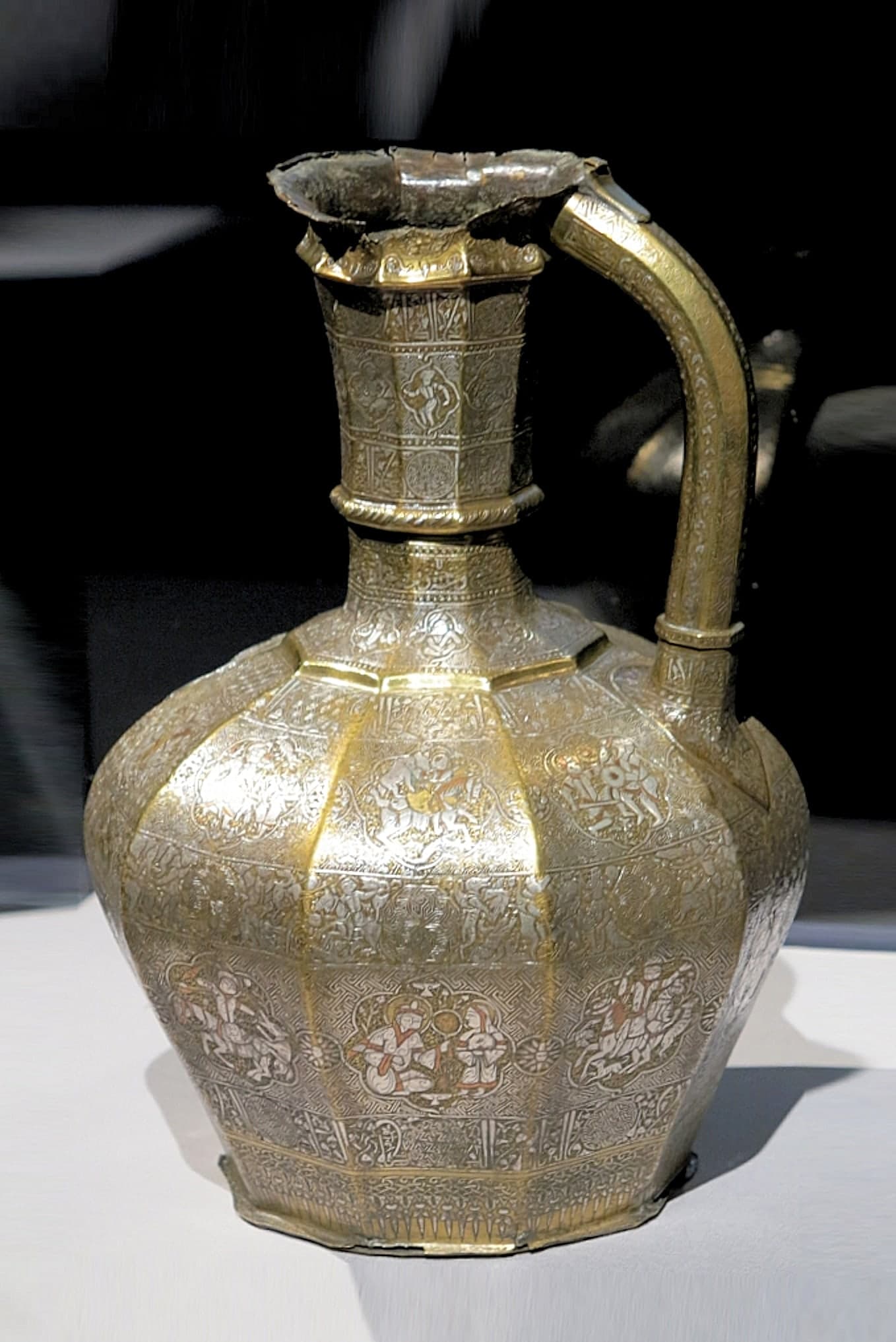 A brass ewer with a pear-shaped body, tall narrow neck, curved handle, and long spout shaped like a bird or dragon head, the surface densely inlaid with silver roundels containing courtly figures and Arabic inscriptions.