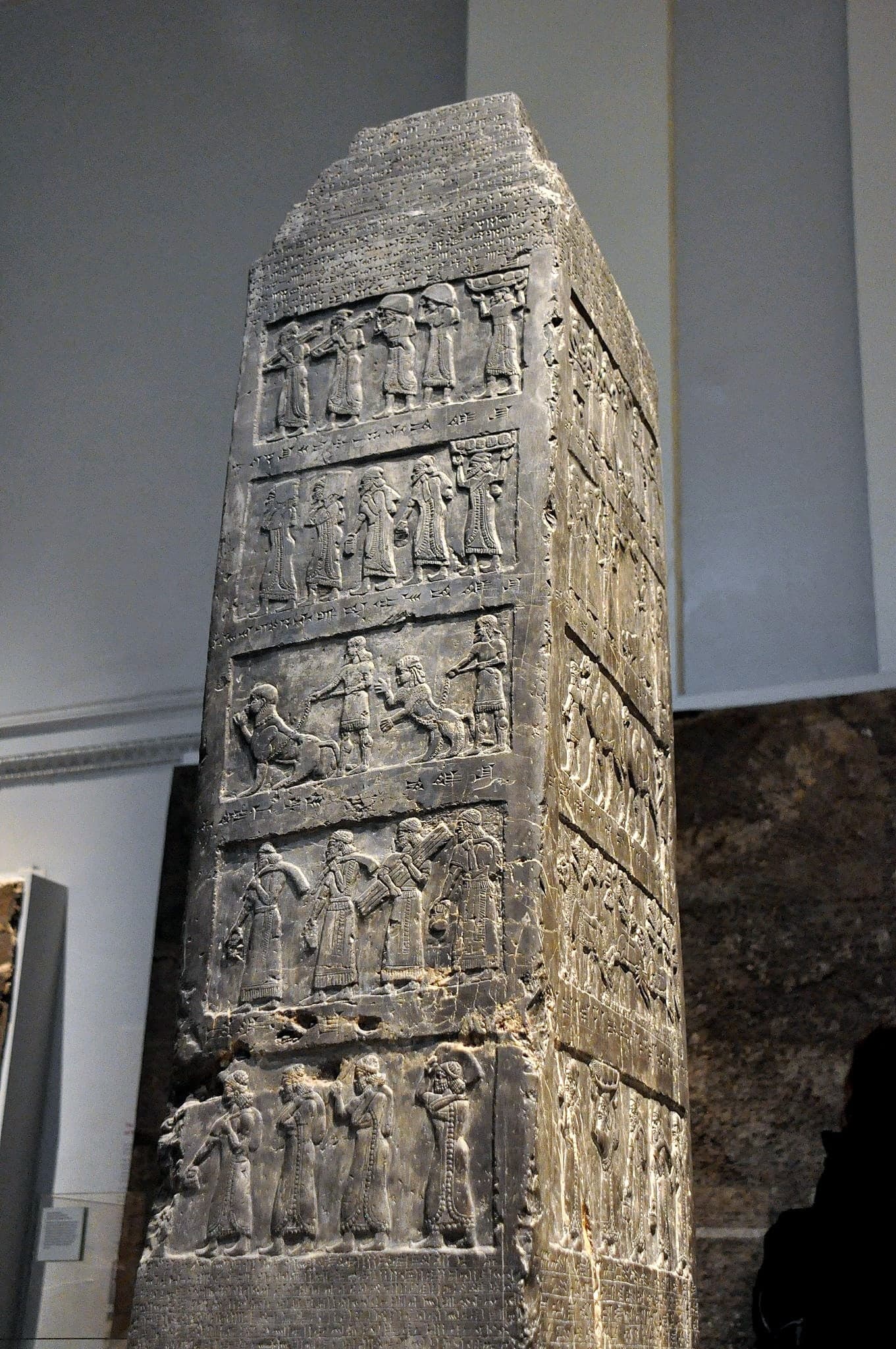 A tall, tapering four-sided shaft of black stone, carved on all faces with horizontal bands of figures and cuneiform text. Each band shows people and animals in profile, bringing goods.