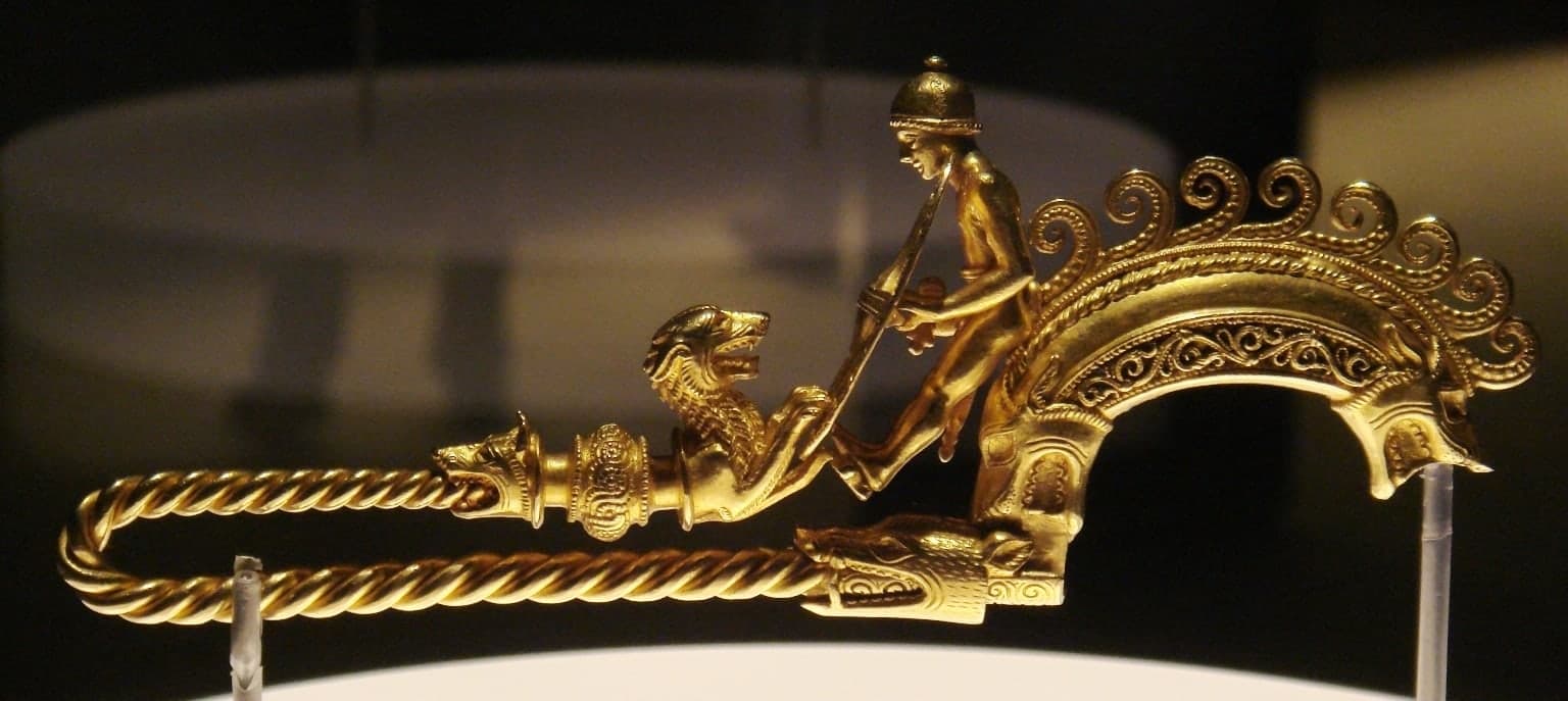 A small gold fibula with glass elements, about five centimetres high and two and a half centimetres wide, shown as a compact ornamental clasp.