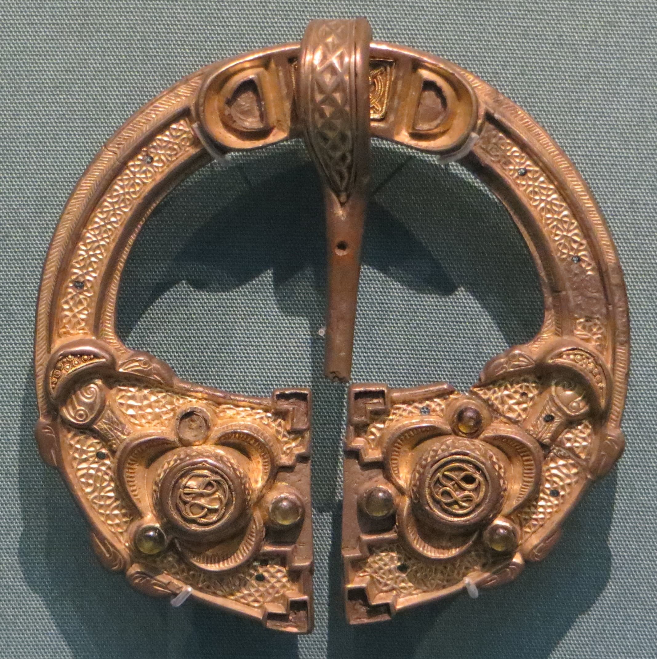 A near-circular silver-gilt brooch, its hoop stopping short of closing to leave a narrow gap, with a long pin lying across the ring. The broadened ends of the hoop carry dense fine ornament and small inset settings; the gilded surface is worn and darkened in places.
