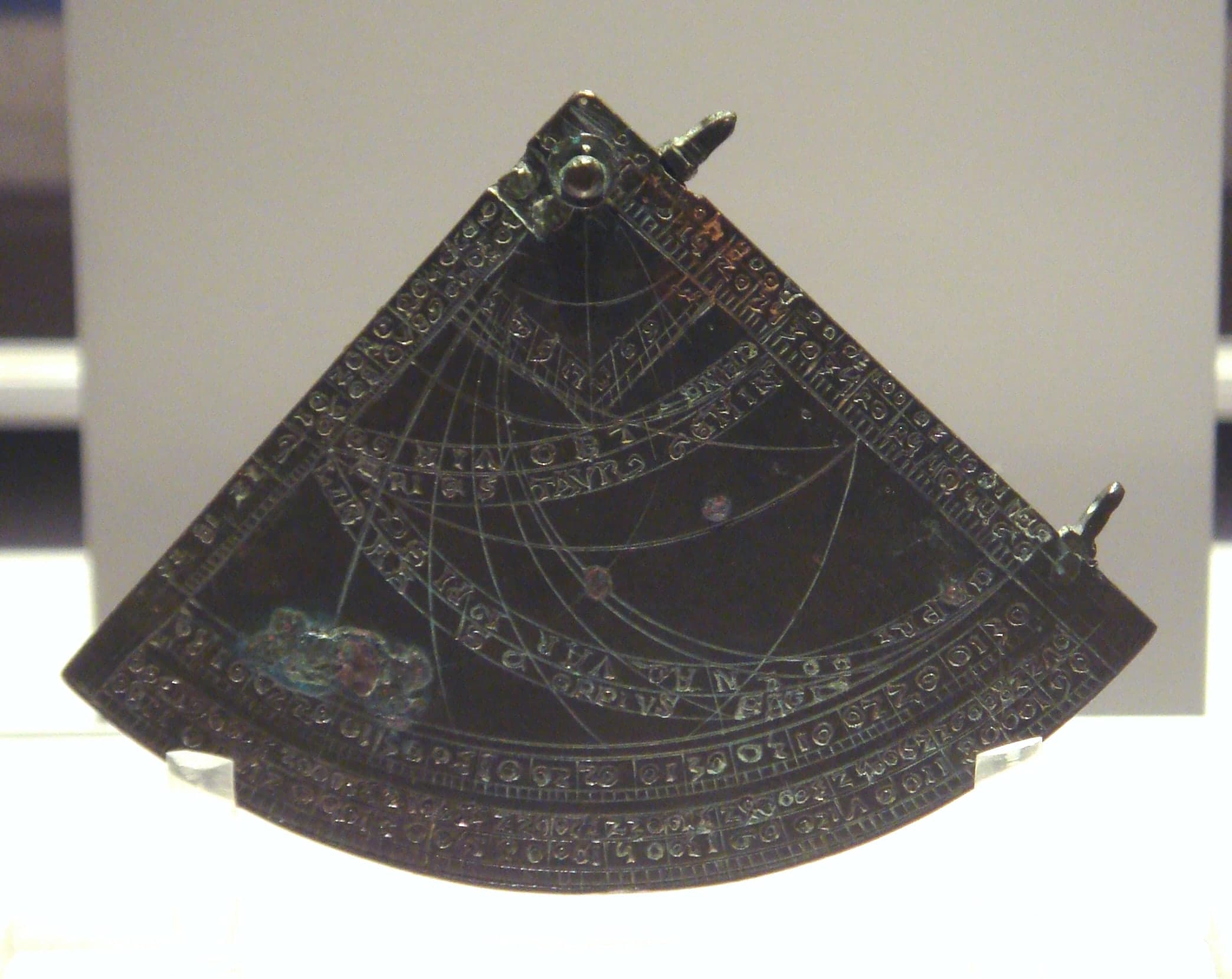 A thin brass quarter-circle engraved with curved lines and arcs, with two small sighting holes along one straight edge.