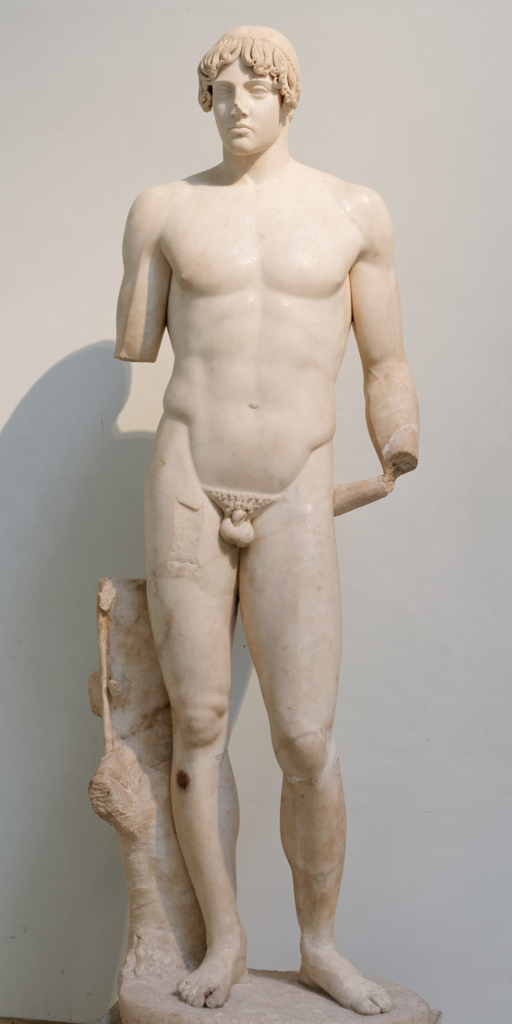A life-size marble figure of a nude youth stands upright. A tree-stump support rises beside the figure’s right side, and a narrow strut connects the left arm and leg.