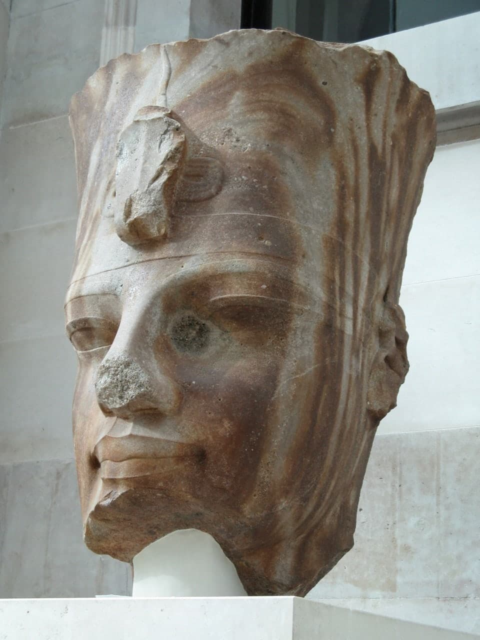 A monumental brown quartzite head of a pharaoh, broken at the neck, with full lips, a broad nose and the remains of a nemes headdress, displayed upright against a plain background.