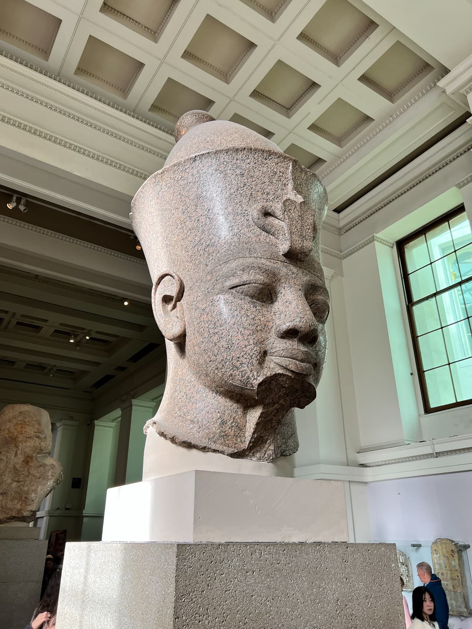 A large, fragmentary granite sculptural head with a separate arm fragment, shown against a pale museum background.