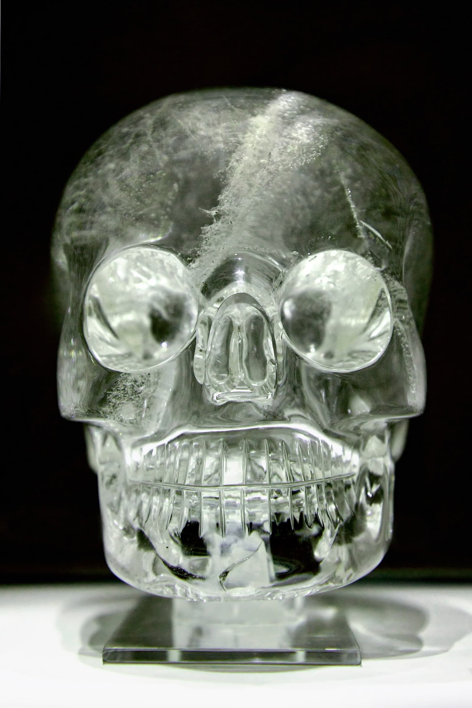 A clear, life-sized human skull carved in translucent crystal, shown facing forward with hollow eye sockets, nasal opening, and a full row of teeth.