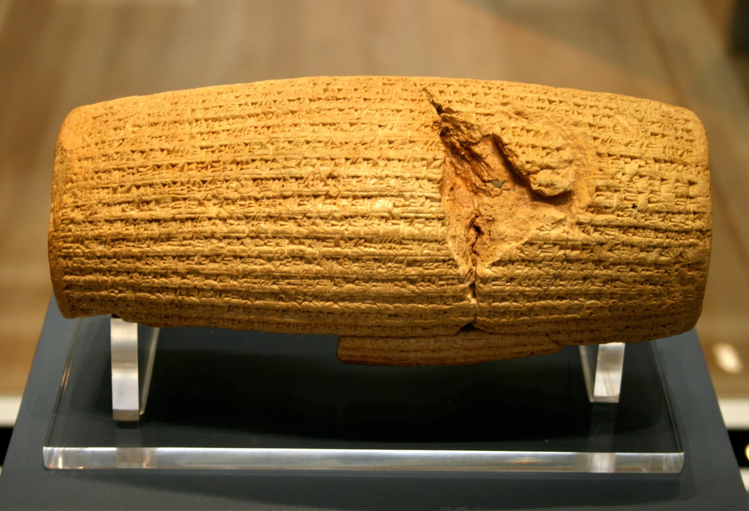 A reconstructed clay cylinder, broken into several pieces and reassembled, covered in neat horizontal lines of cuneiform script running around its curved surface.