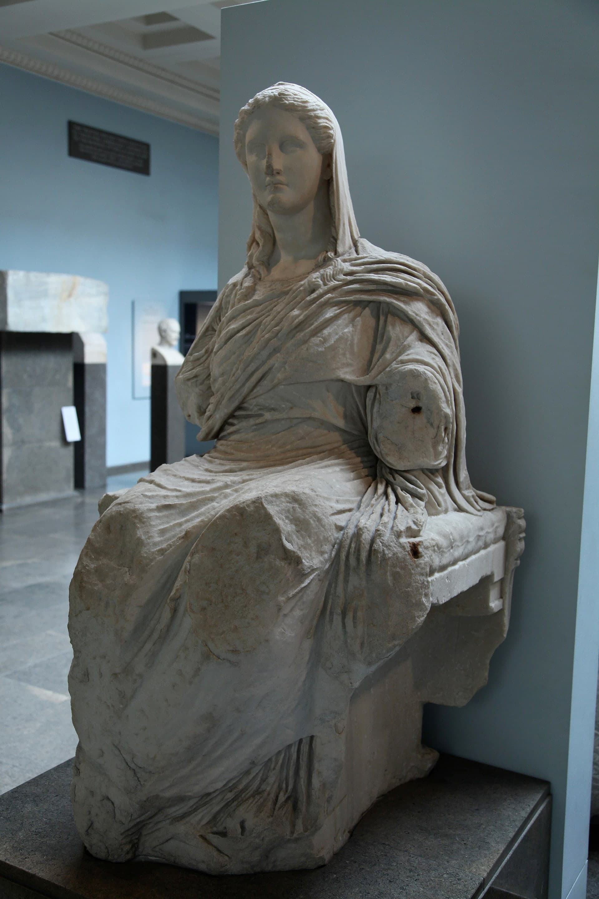 A seated marble figure, missing both arms below the shoulder, draped in carved cloth that falls in folds across the lap and legs. The head is intact, facing forward, with a calm expression.