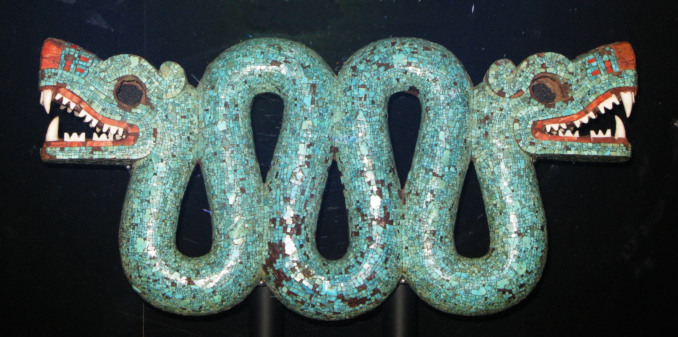 A wooden serpent covered in turquoise mosaic, curved into a shallow S-shape with a head at each end. The mouths gape, showing white shell teeth, and a single red forked tongue emerges from the centre.
