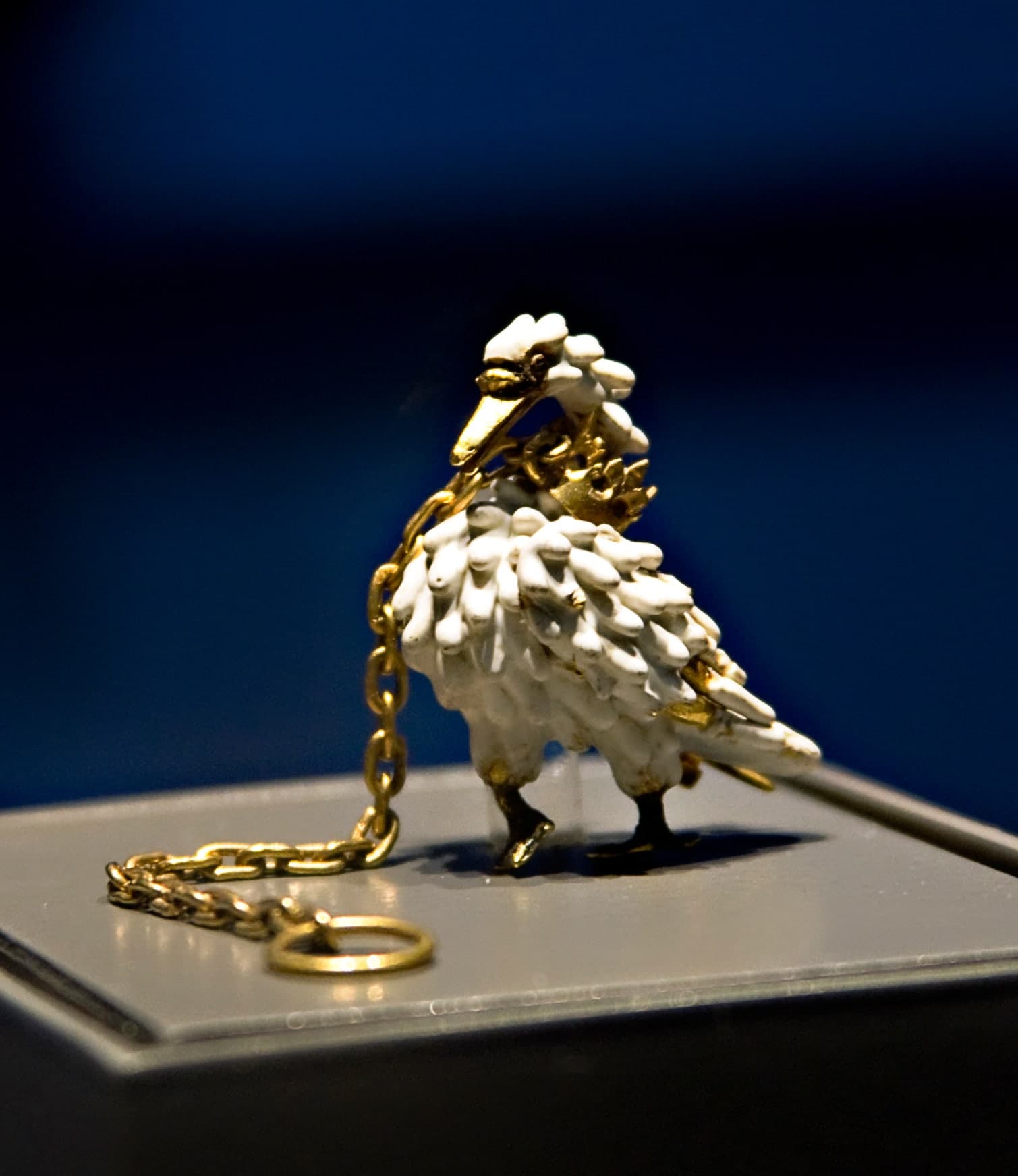 A gold and enamel brooch in the shape of a standing or walking mute swan. The swan is collared by a gold crown with six fleur-de-lys tines, and has a pin on its right side for fastening. A gold chain ending in a ring is attached to the crown.