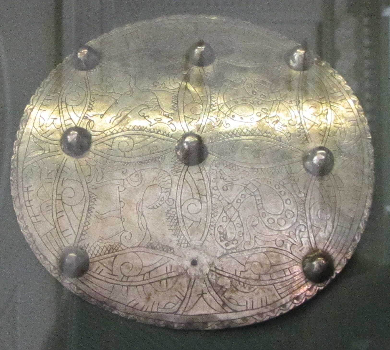 A circular silver brooch with conical bosses and dense engraved animal and plant ornament in overlapping circles; one boss is missing from the arrangement.