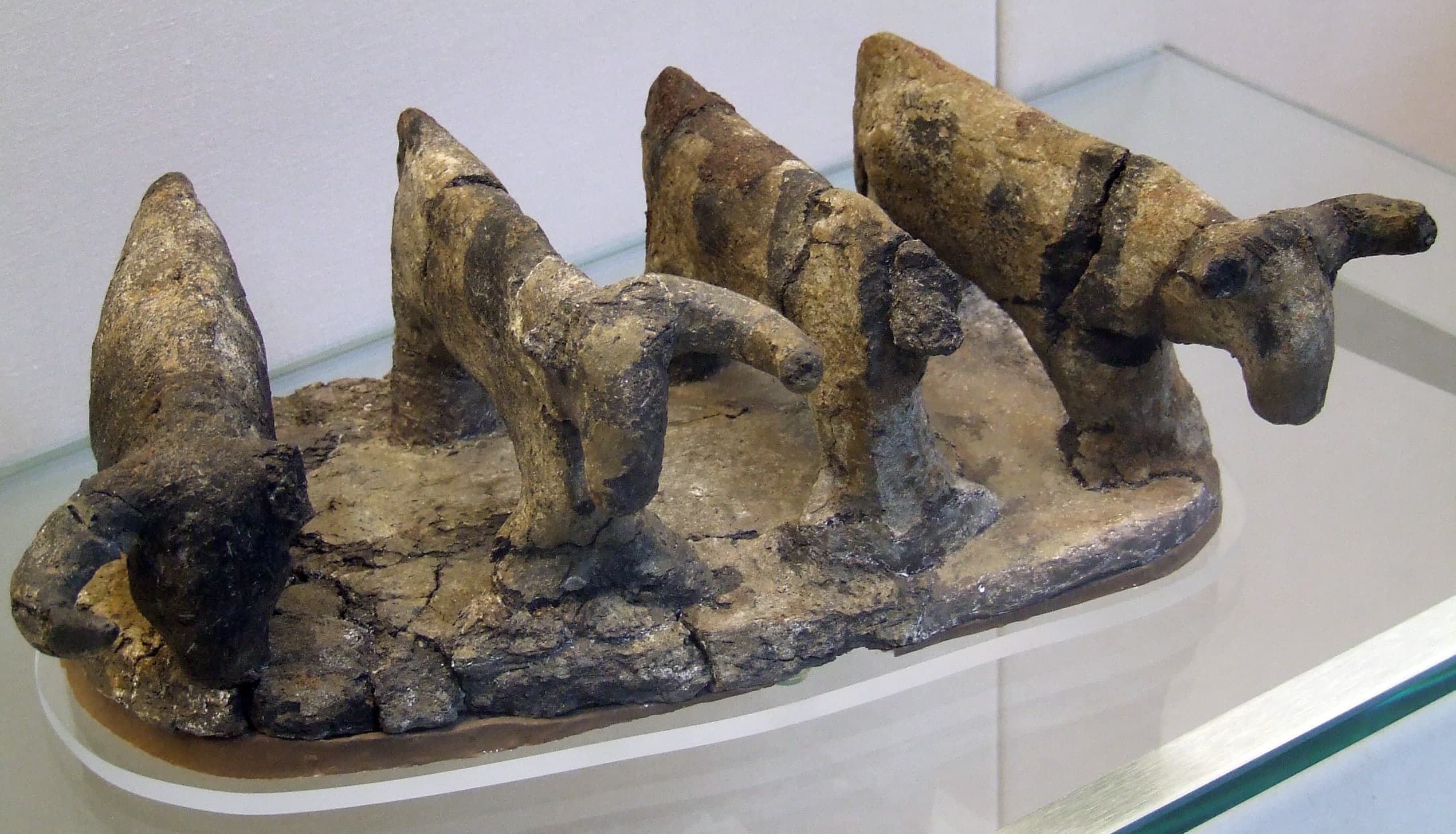 Four small clay cattle standing in a row, horns curving inward and down; one head missing, paint largely worn away, on a plain base.