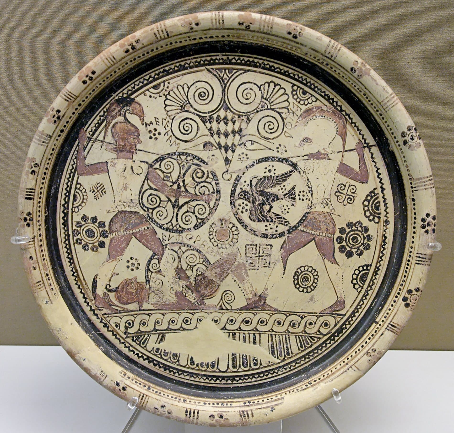 A circular ceramic plate painted with three figures: two standing warriors with spears and shields facing each other across a horizontal body on the ground, with painted letters labelling each figure.