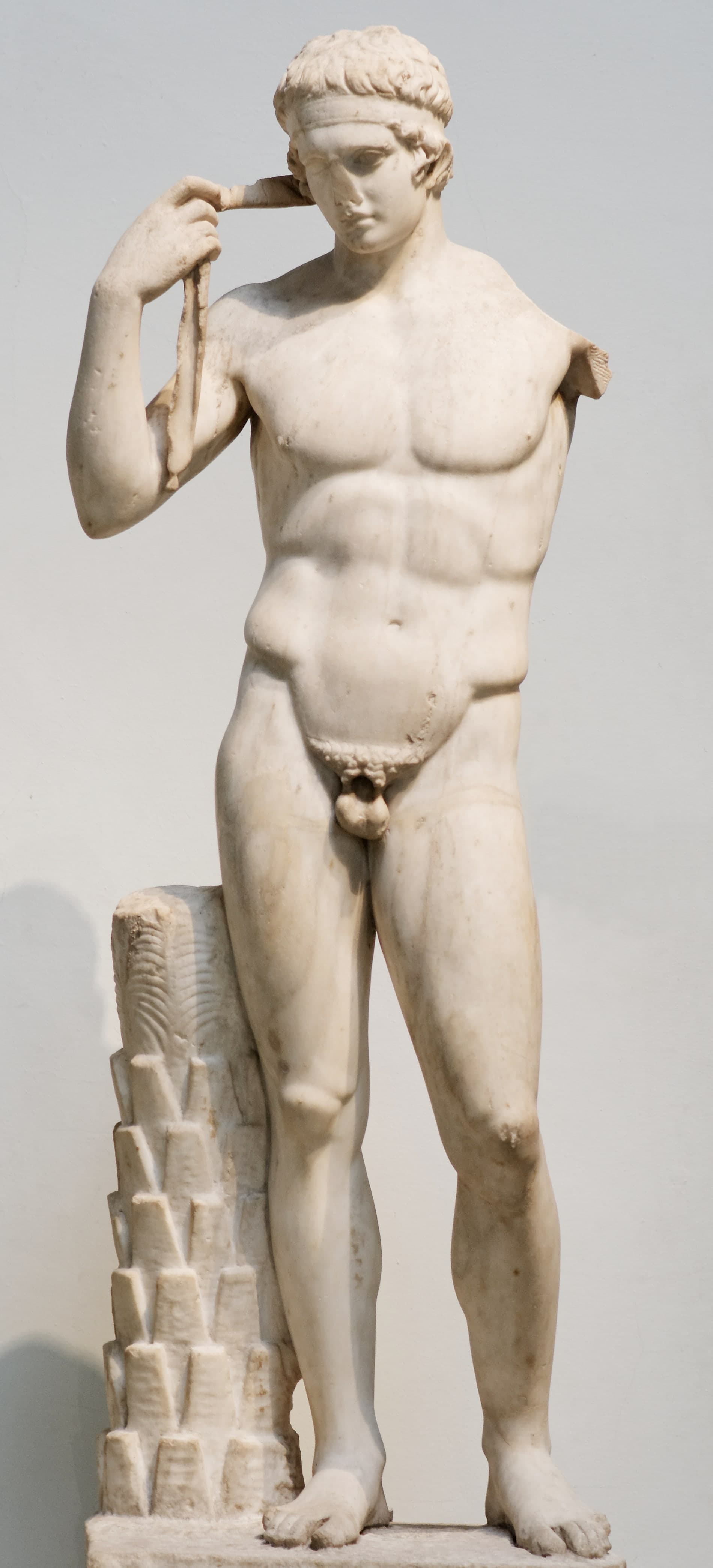A pale marble statue of a nude youth standing with both arms raised as if tying a narrow band around his head, weight shifted onto one leg in a balanced athletic pose.
