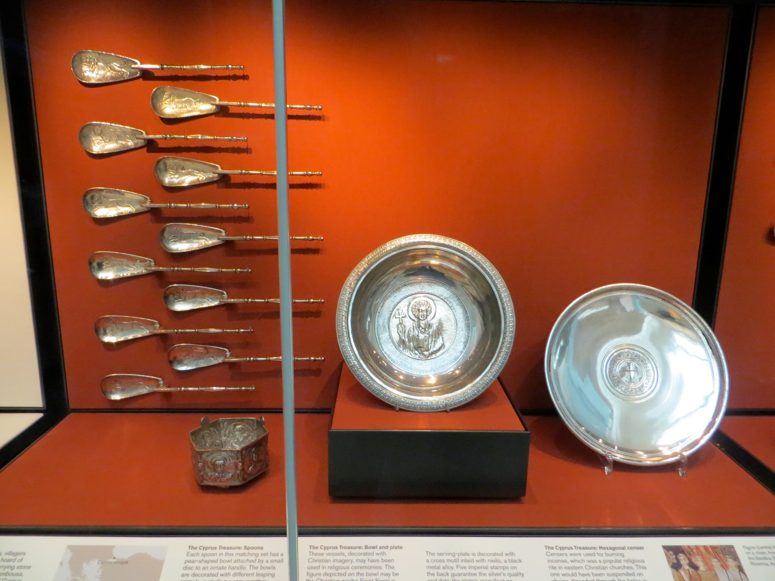 A display case containing twenty-eight silver artifacts arranged in rows. The items include plates, chalices, cups, and bowls, some plain and others intricately designed with crosses or other symbols.
