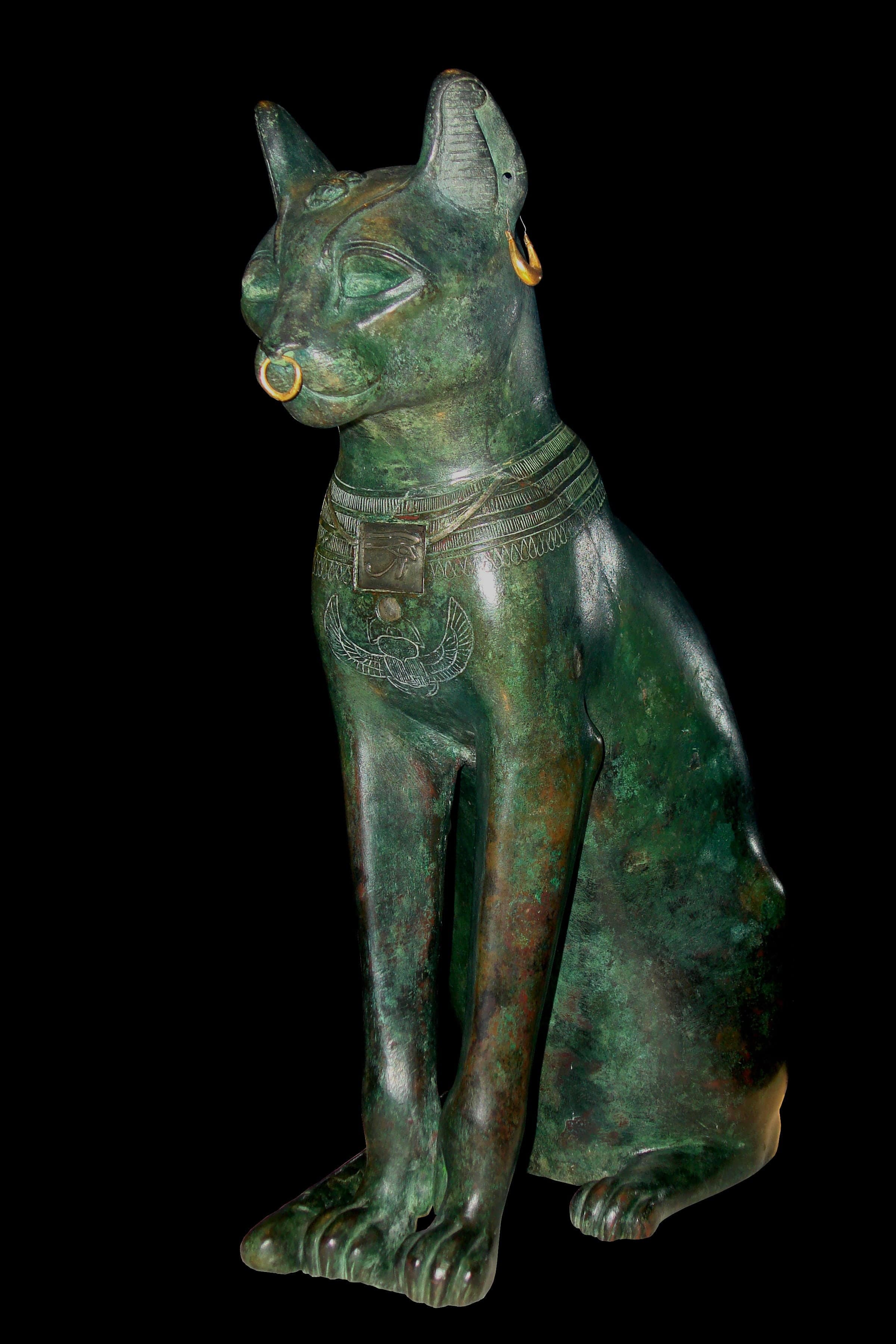 A seated bronze cat with upright posture, ears forward, tail wrapped around its front paws. Silver earrings hang from pierced ears, and a gold necklace with a pendant rests at its chest.