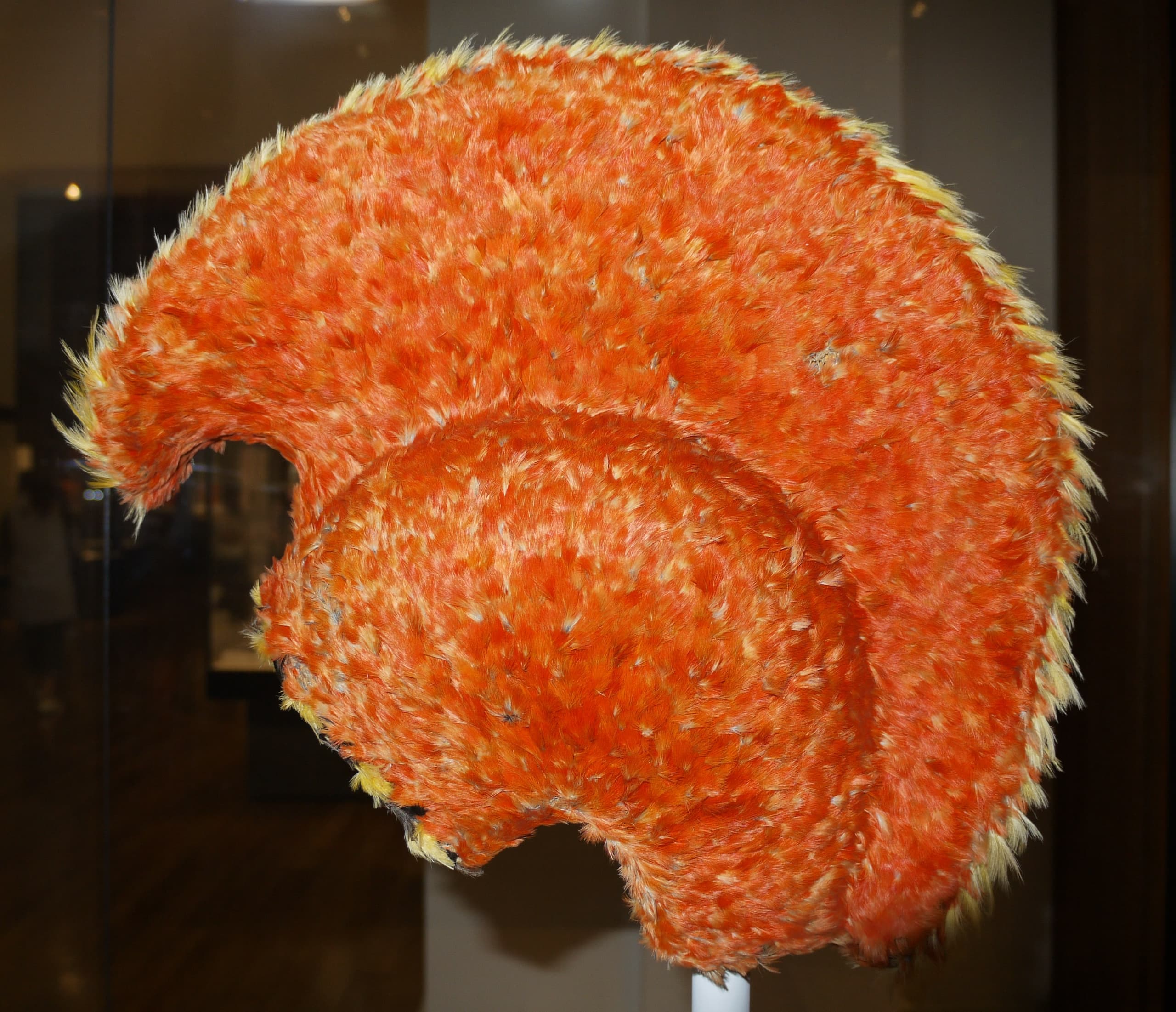A crested helmet covered entirely in small feathers, woven onto a shaped frame, with a smooth curved surface that rises to a point at the back.