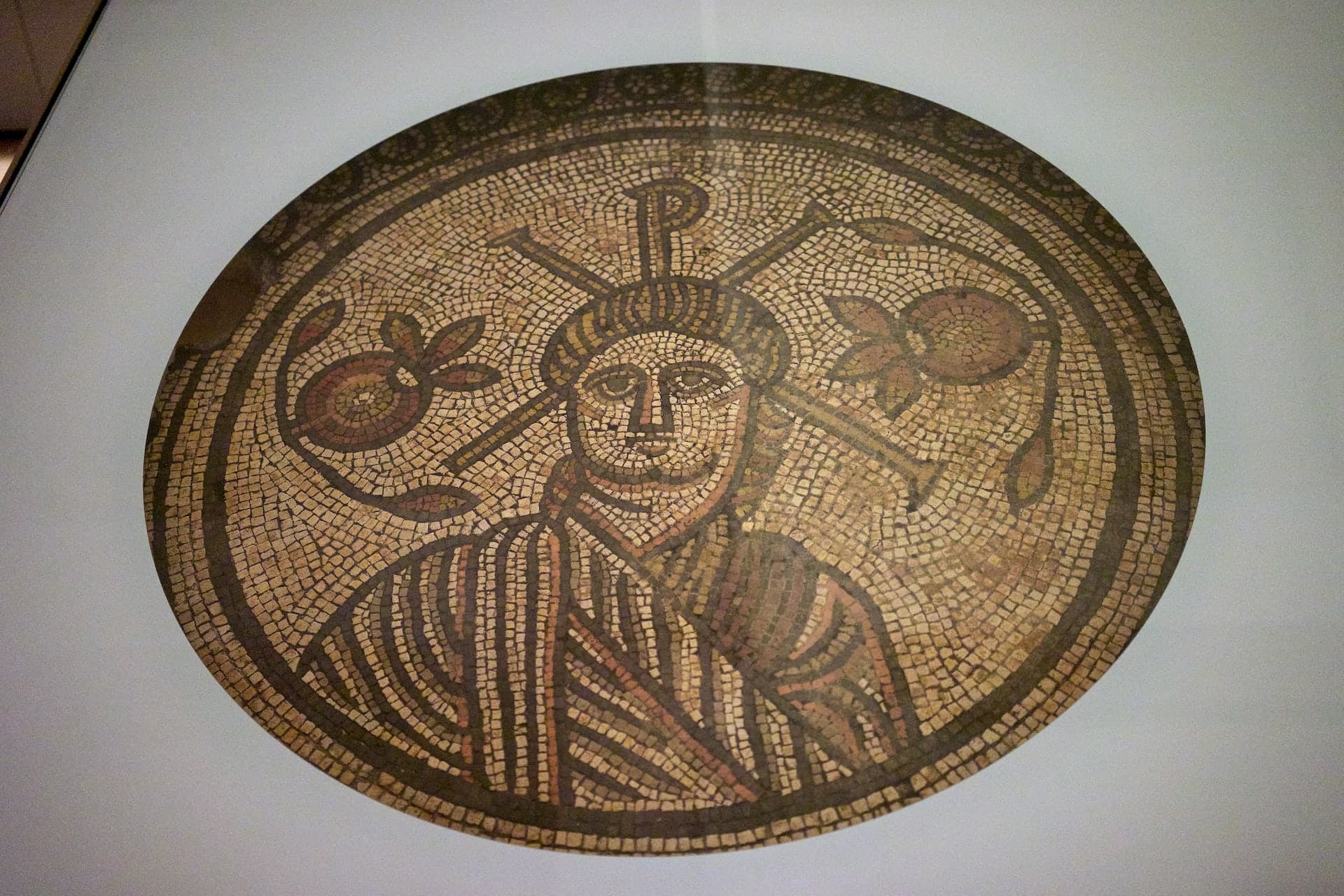 A circular mosaic panel of small stone cubes in cream, dull red and yellow, showing a human head and shoulders facing forward within decorated borders. Old breaks and patches cross the surface.