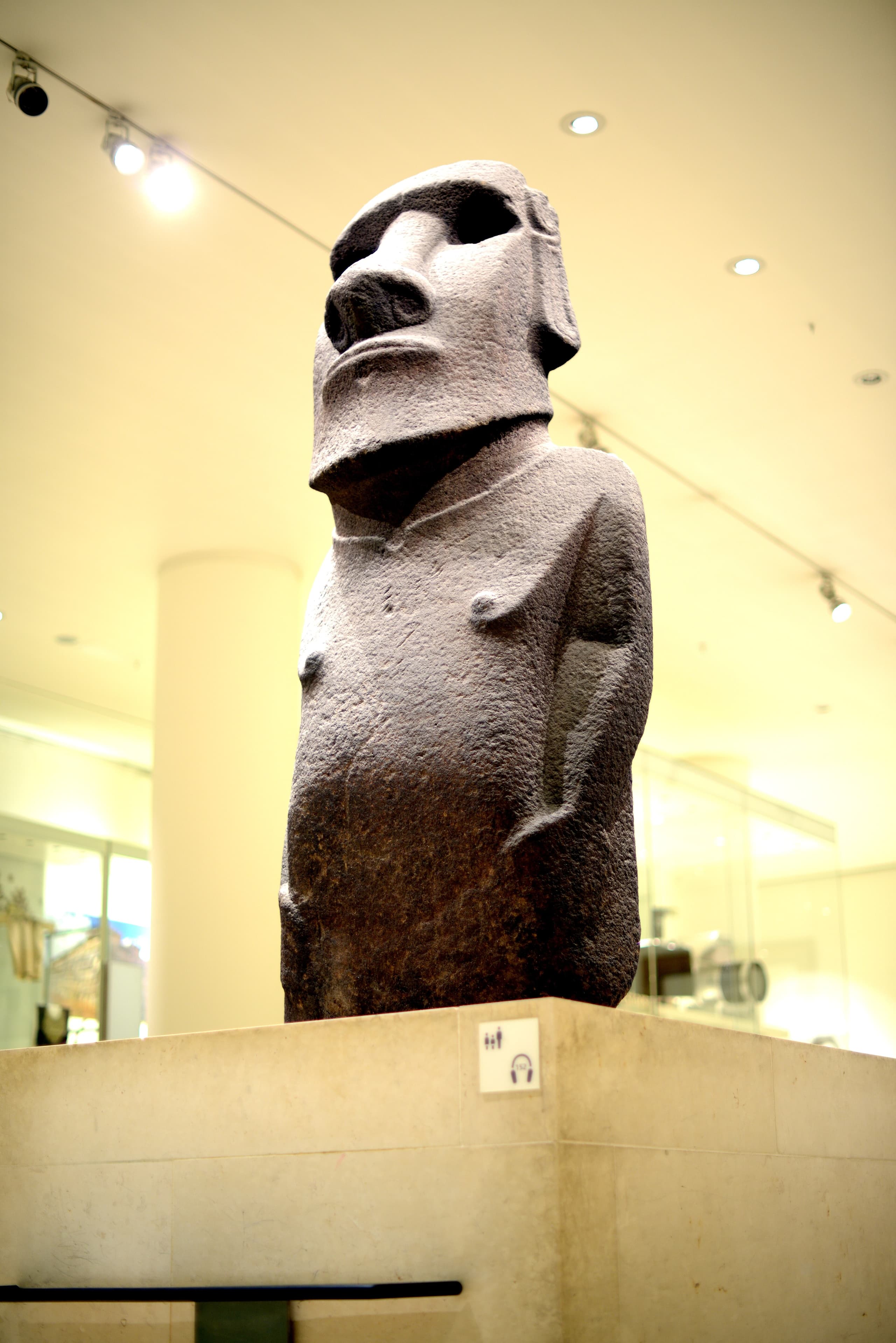 A dark grey basalt statue of a standing human figure, upright and stylised, with a large head, prominent brow, long nose, and arms at the sides, hands meeting at the abdomen.