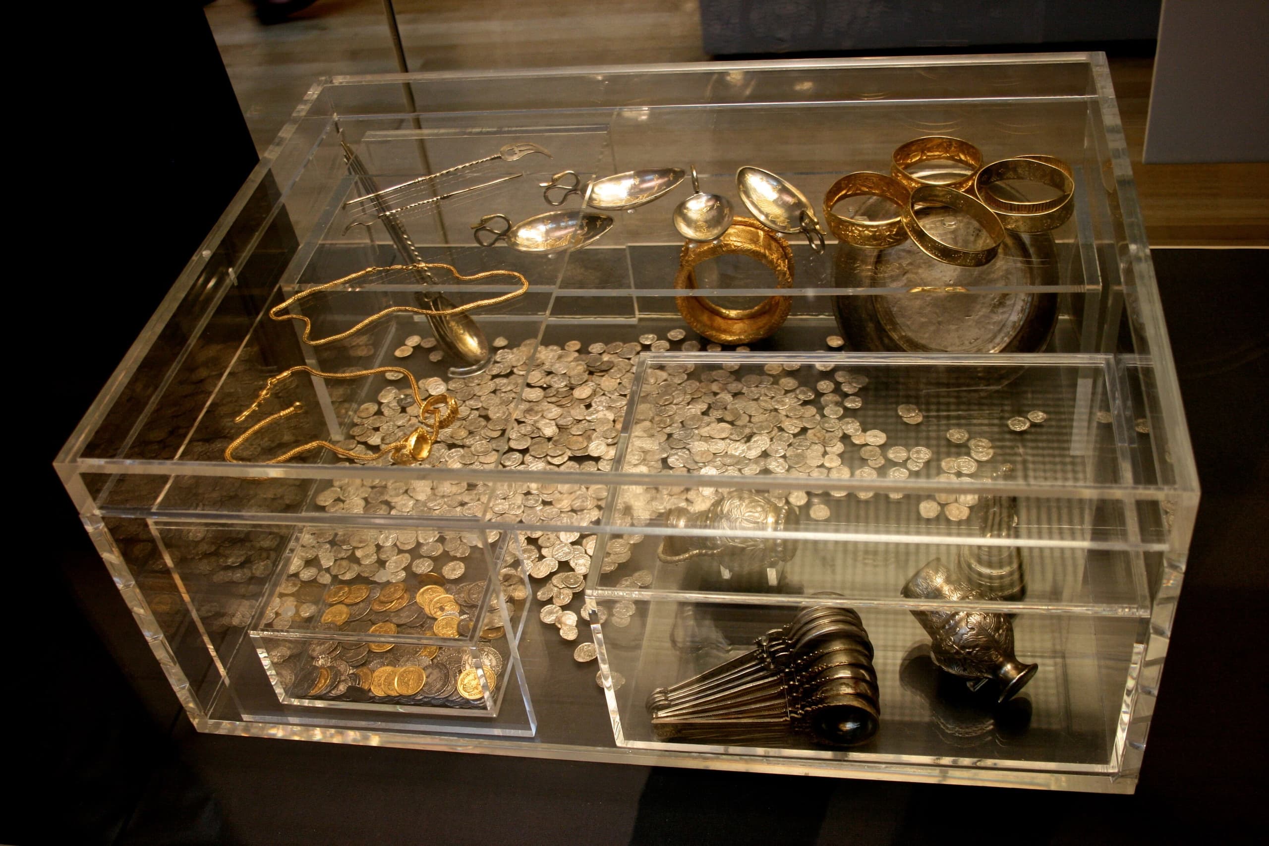 Gold jewellery, silver tableware and dense stacks of late-Roman coins arranged as a hoard: small worked vessels, a body-chain and coin piles against a neutral display ground.