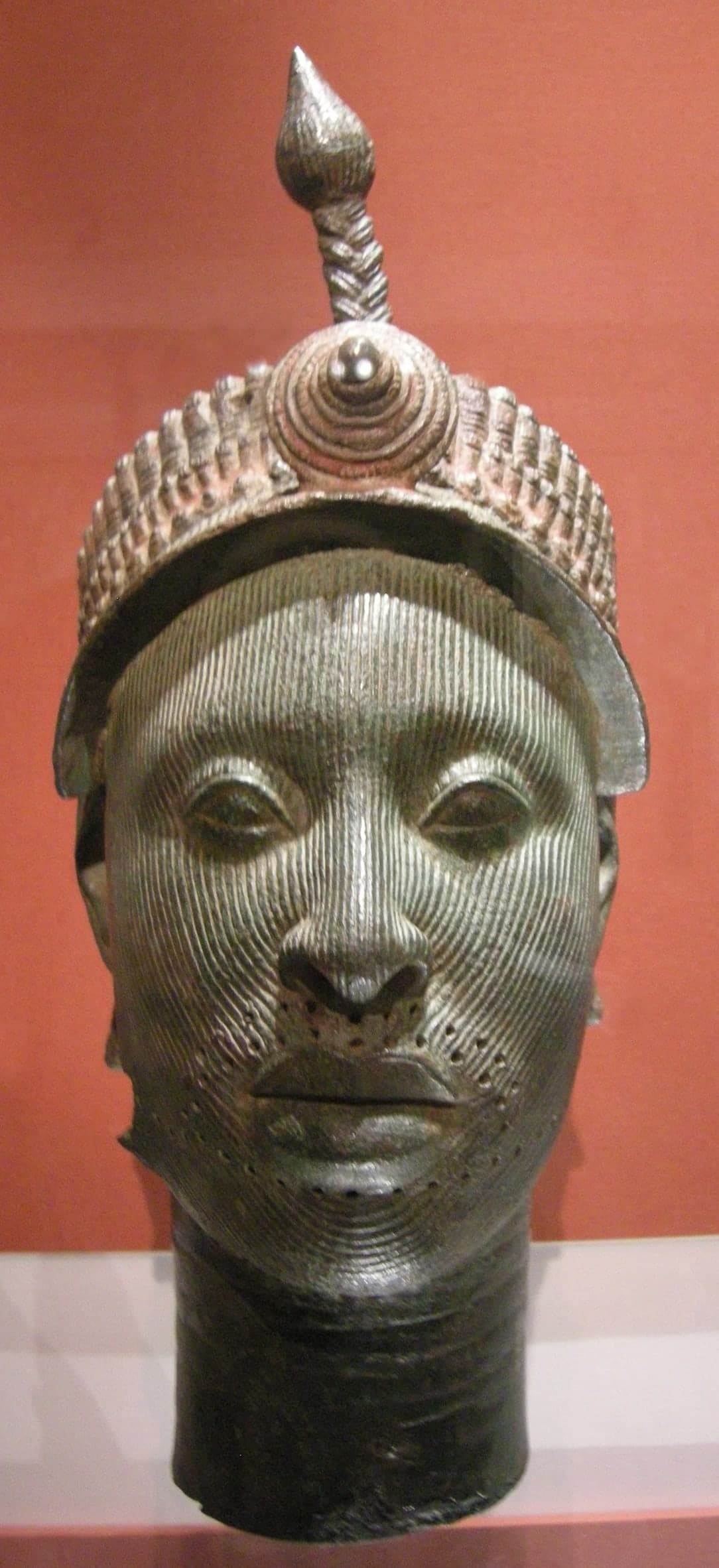A life-size brass head with idealised features, parallel vertical striations across the face, and small holes around the hairline and jawline where attachments would have been fixed.