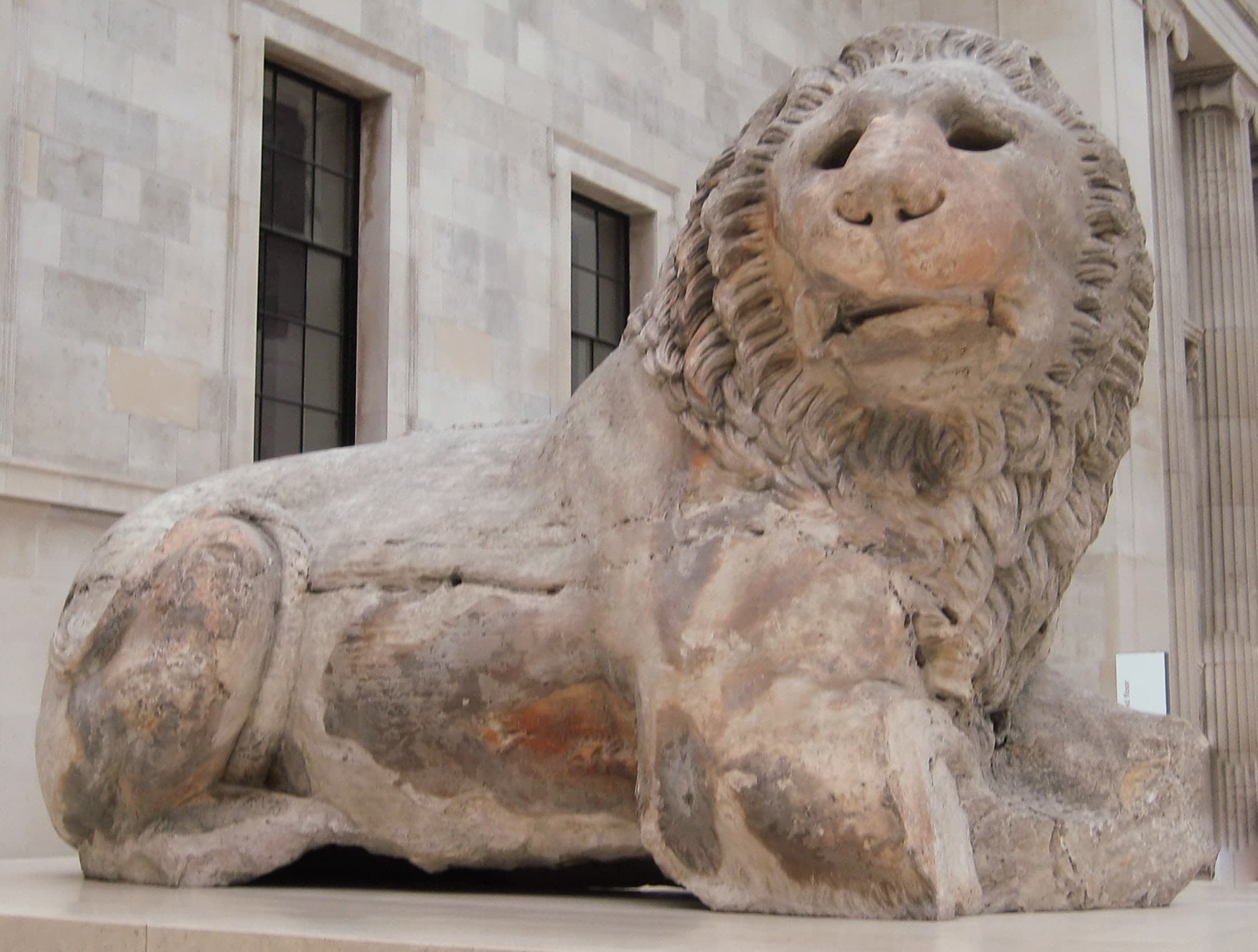 A large marble lion seated on its haunches, front legs extended, head raised. The surface is weathered and details softened by exposure.