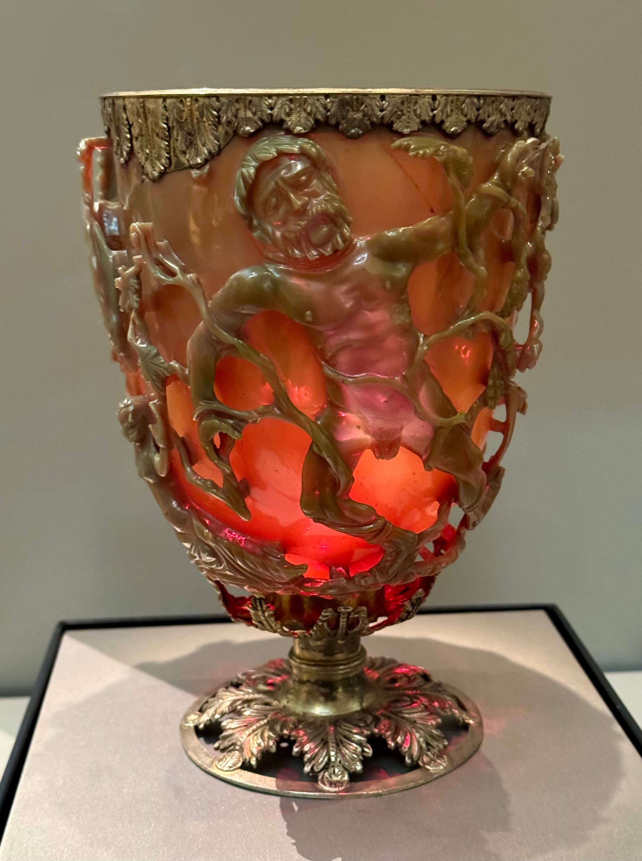 A cup carved from dark glass with openwork figures and vines forming a cage around the vessel. The glass appears greenish in the photograph, with the carved scene showing human figures and foliage in relief.