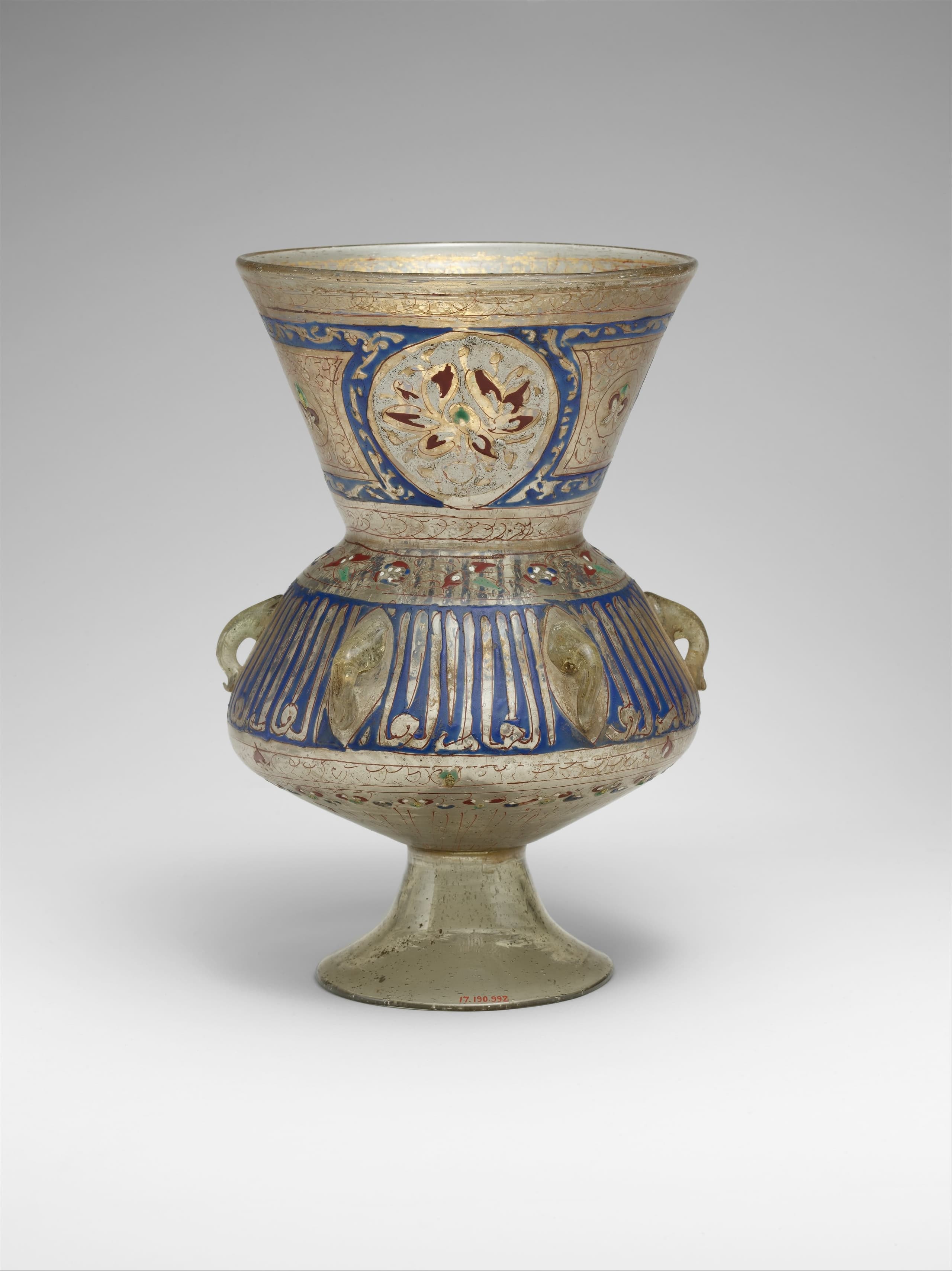 A bulbous glass vessel with a flared neck, decorated with bands of Arabic calligraphy in blue and gold enamel, with small loops projecting from the sides for suspension chains.