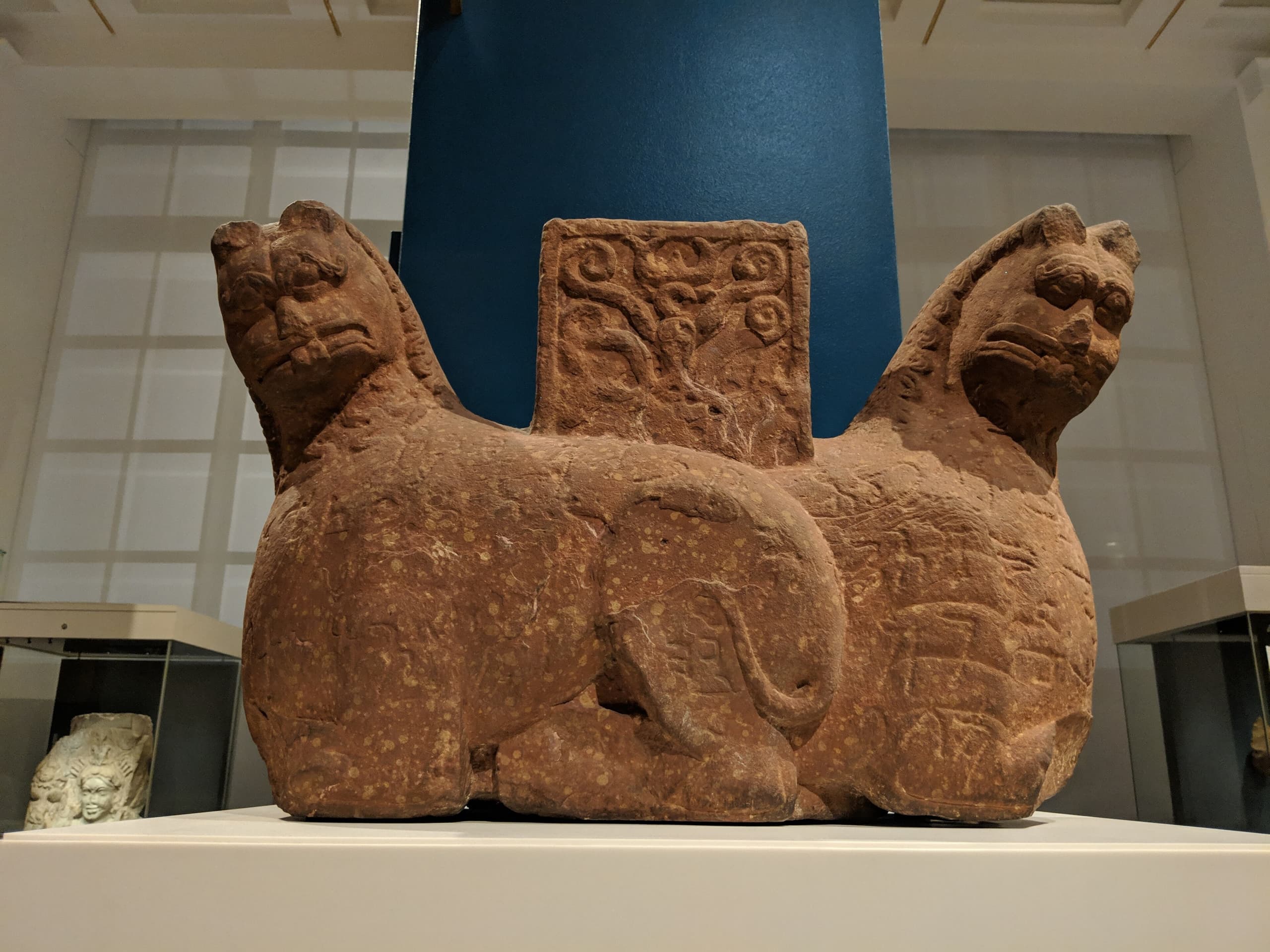A sandstone capital carved as two lions back to back, their bodies dense with small incised lettering, a Buddhist triratana emblem set between them at the centre.