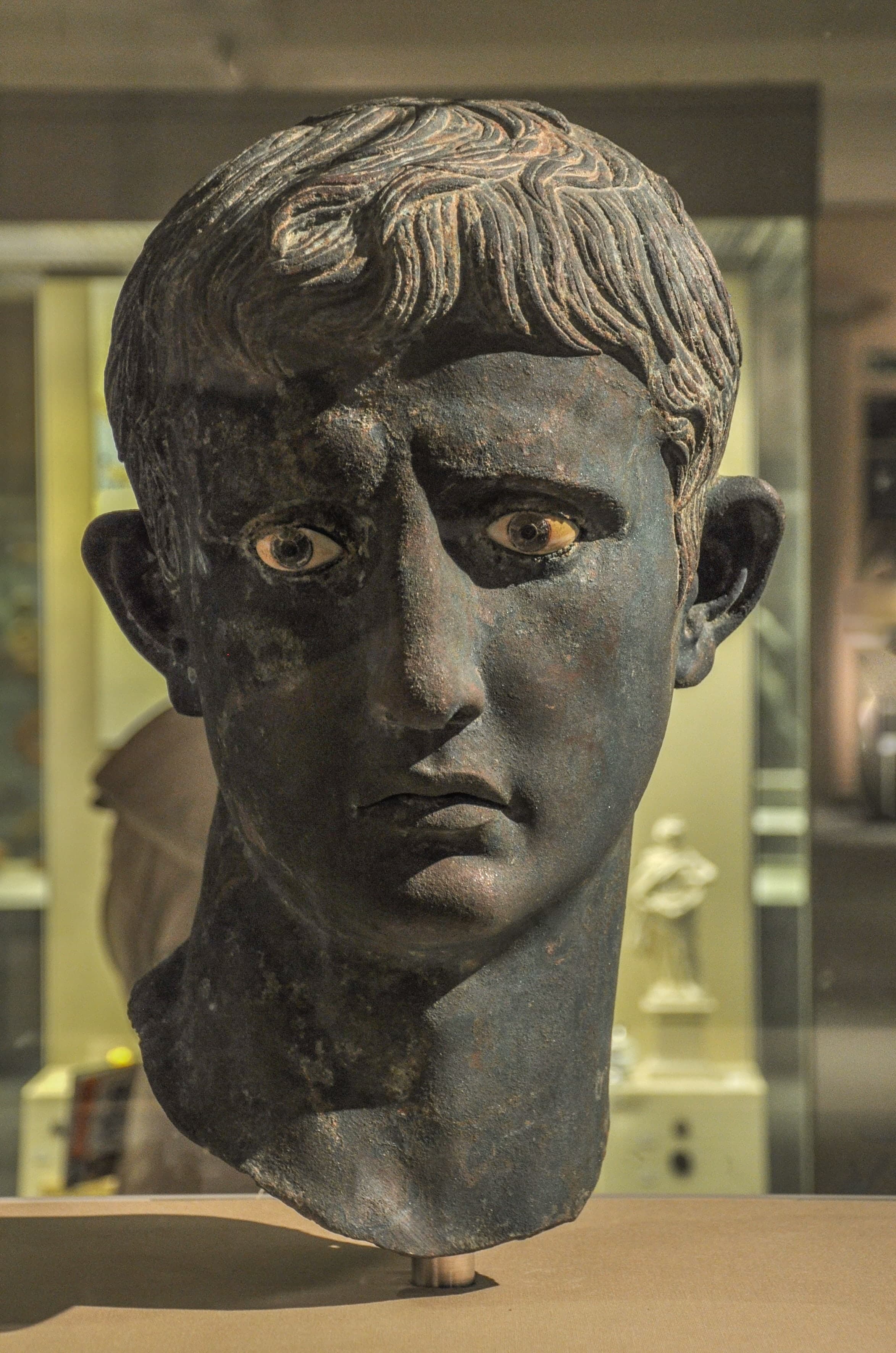 A bronze head of a young man with short hair combed forward, idealised features, and inlaid eyes of white calcite and glass. The neck is broken off roughly below the jawline.