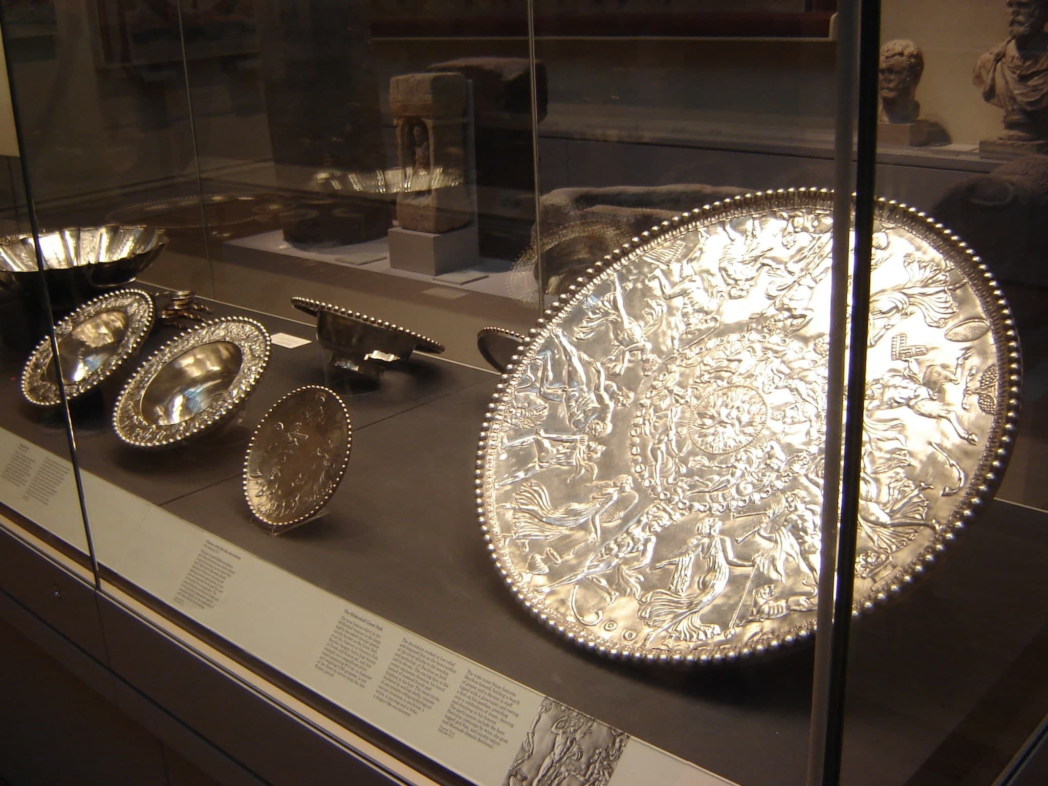 A large circular silver dish with a bearded face at the centre, surrounded by a frieze of human and animal figures in relief, with a decorative border of scallop shells around the rim.