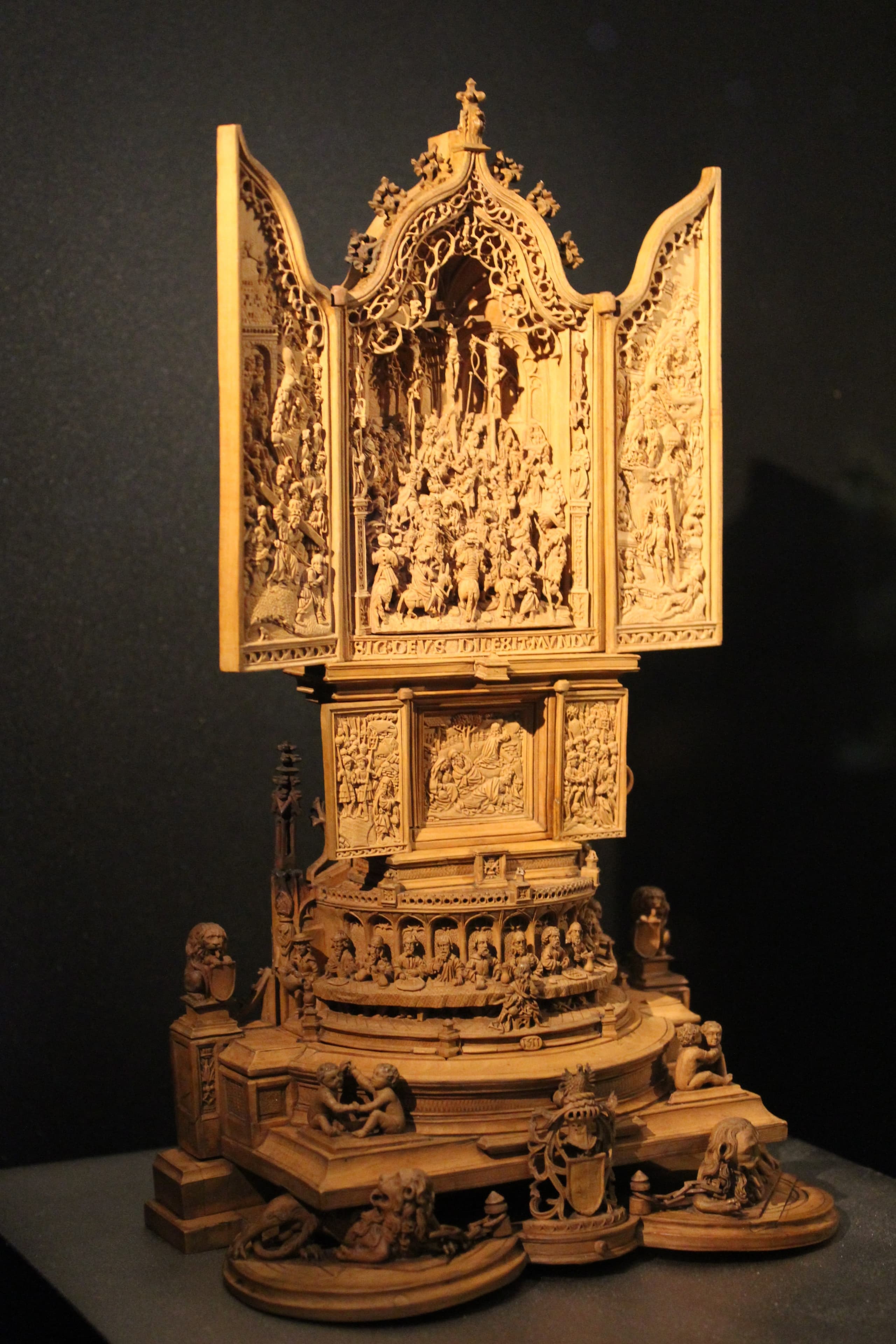 A small carved altarpiece described as a tiered architectural structure, with opening triptych wings, relief scenes from the Passion, arcades and stacked panels.