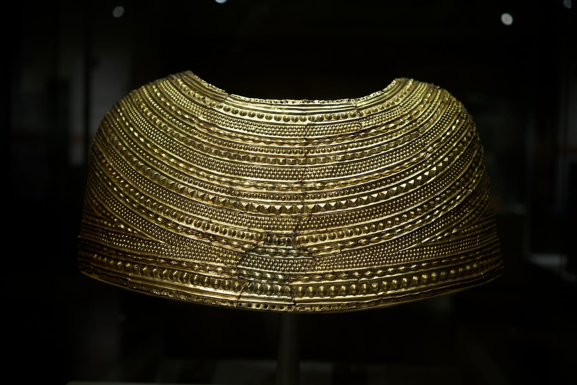 A crescent-shaped gold cape, heavily decorated with concentric bands of raised ribs and small circular bosses, fitted to cover the shoulders and upper chest with small armholes.