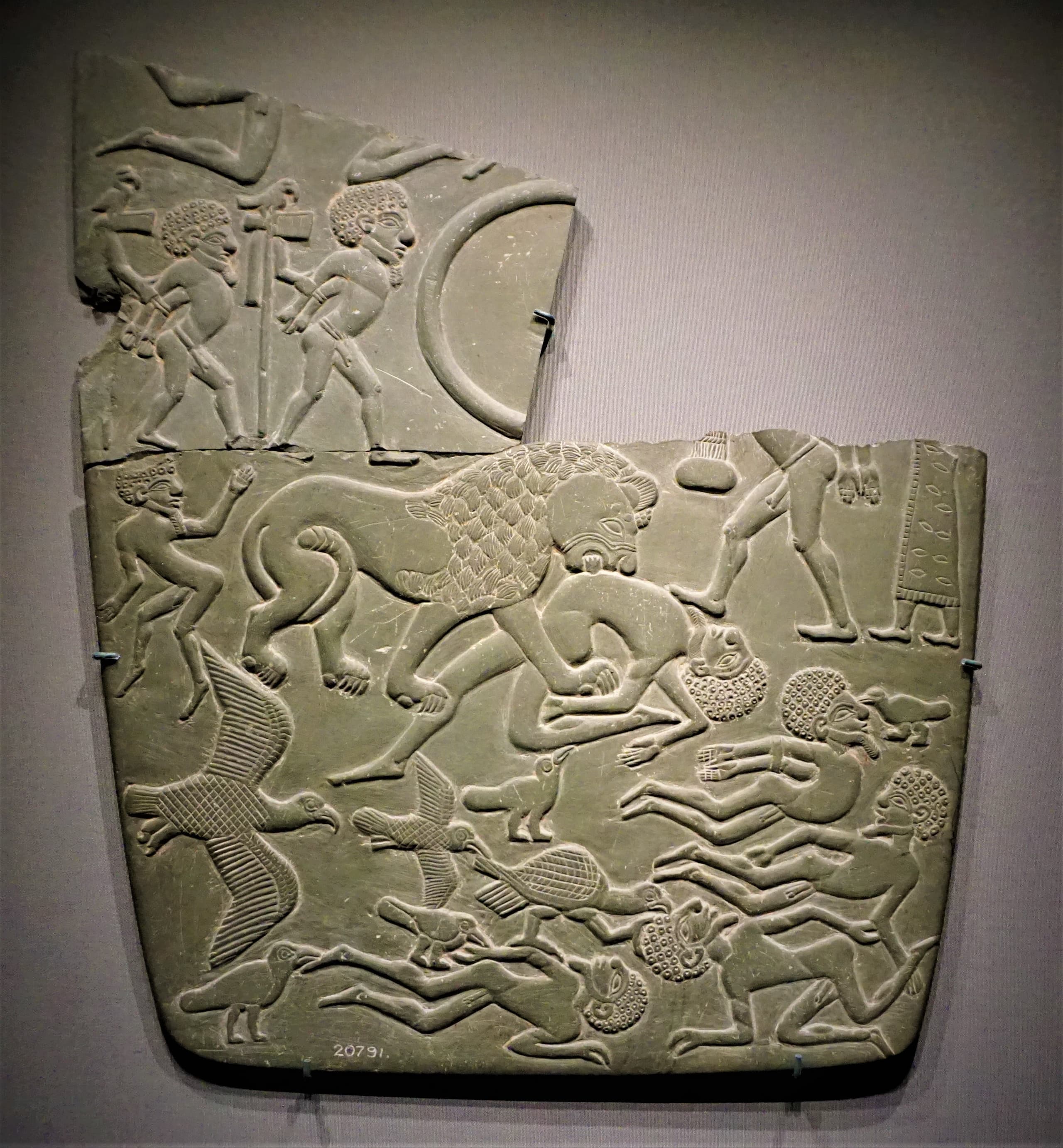 Fragment of a dark grey mudstone palette carved in low relief with human figures, animals, and vertical standards topped with emblems.