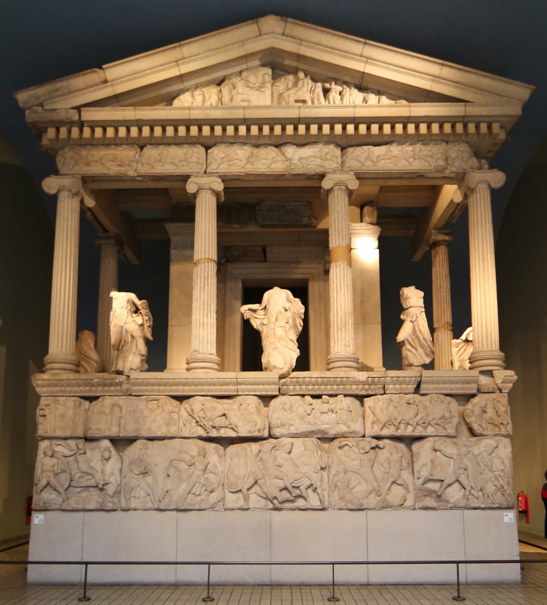 Reconstructed stone façade of a temple-like structure with columns, carved female figures between the columns, and sculpted friezes on the base, displayed inside a museum gallery.