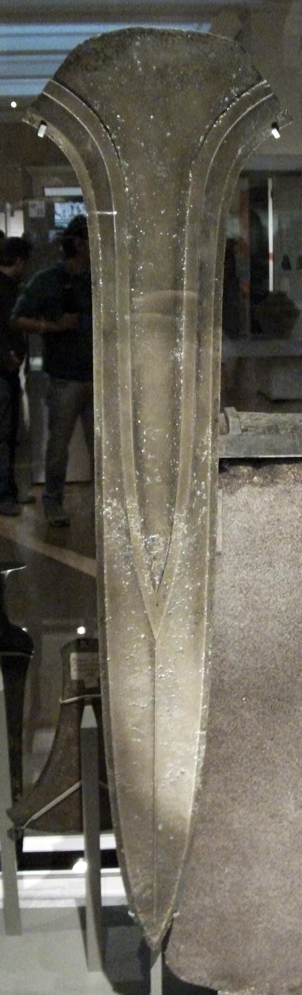A large, dark dirk with a broad blade and heavy section, far wider and thicker than an everyday thrusting weapon, shown as a prehistoric museum object.