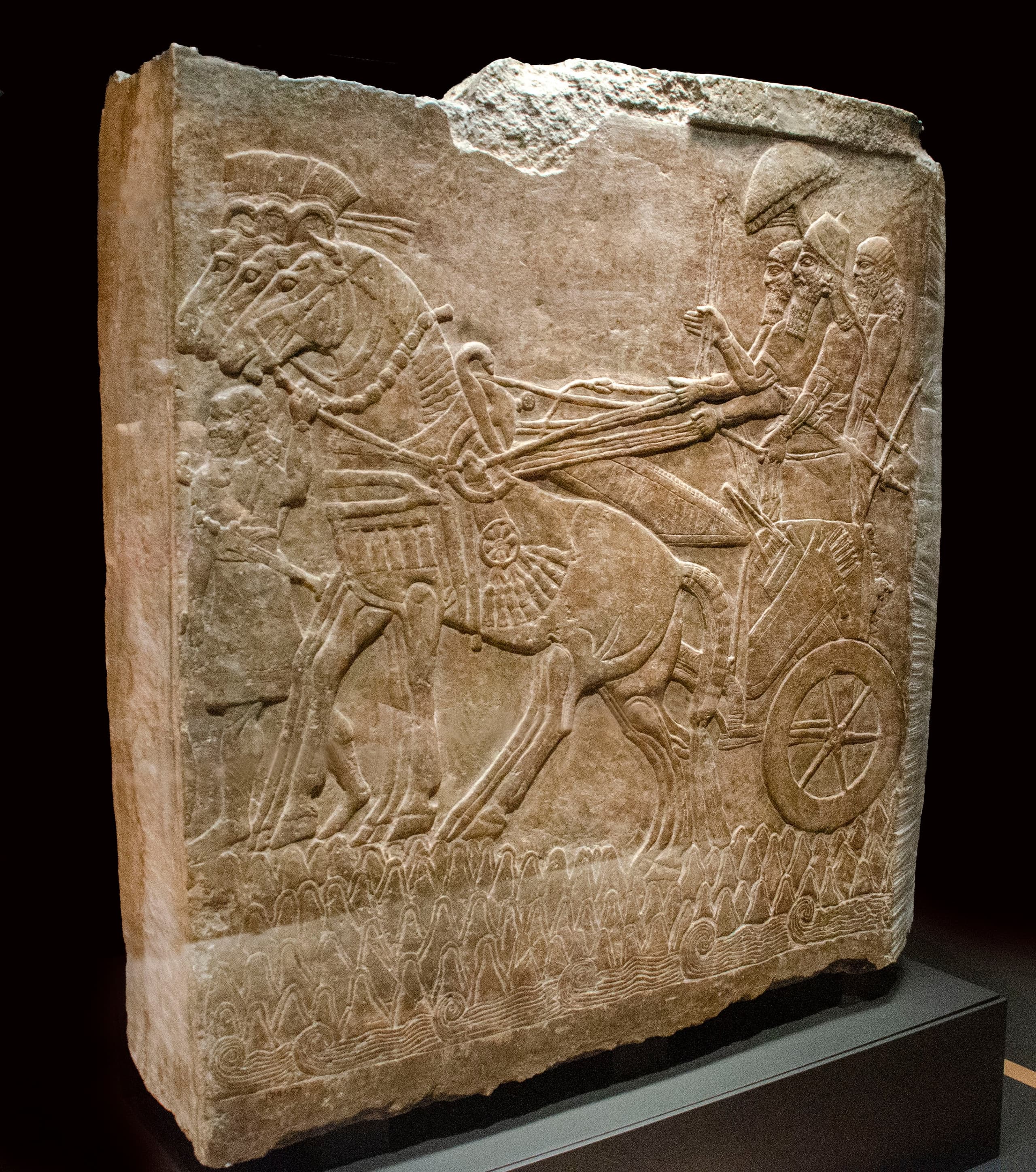 A shallow relief carved in pale stone showing a bearded, winged figure in profile, holding a small bucket in one hand and raising a cone-shaped object in the other.