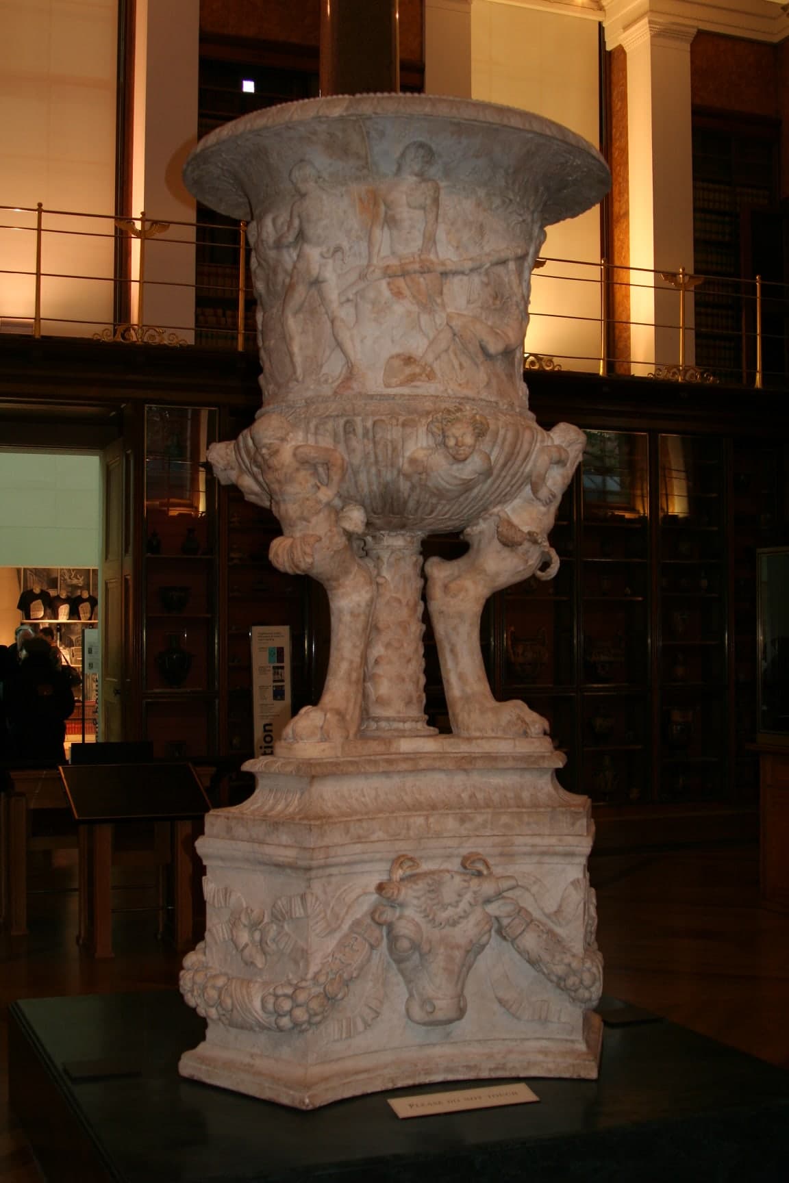 A large marble vase standing on three legs with a triangular base, featuring intricate reliefs around its sides depicting a scene of satyrs making wine. The upper part is inspired by the Borghese Vase, while the lower section is influenced by the Torlonia Vase.