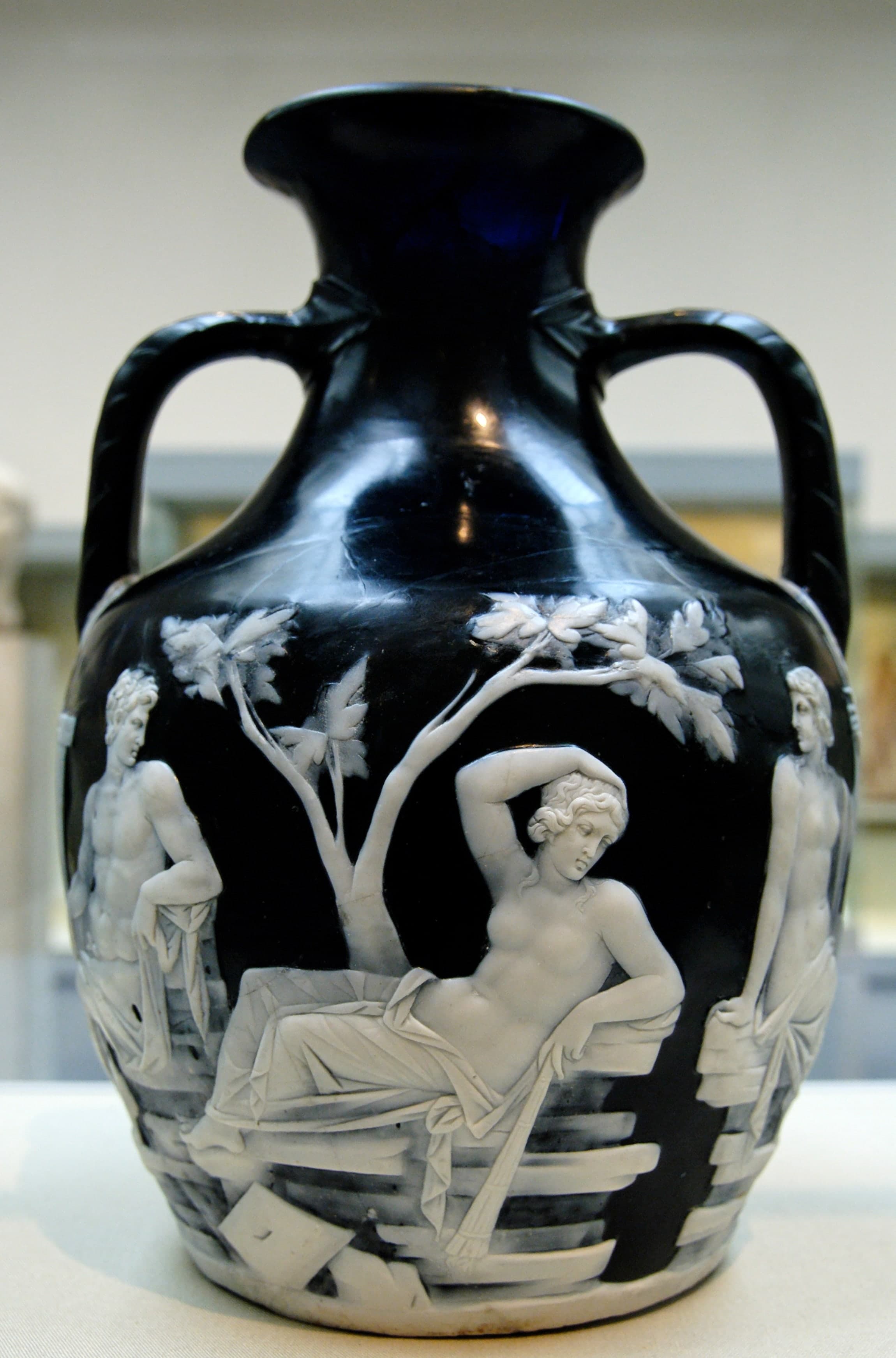 A dark blue glass vase with two handles, its surface carved with white figures in relief: seven human figures, a snake, and bearded heads beneath the handles, arranged in a continuous scene around the body.
