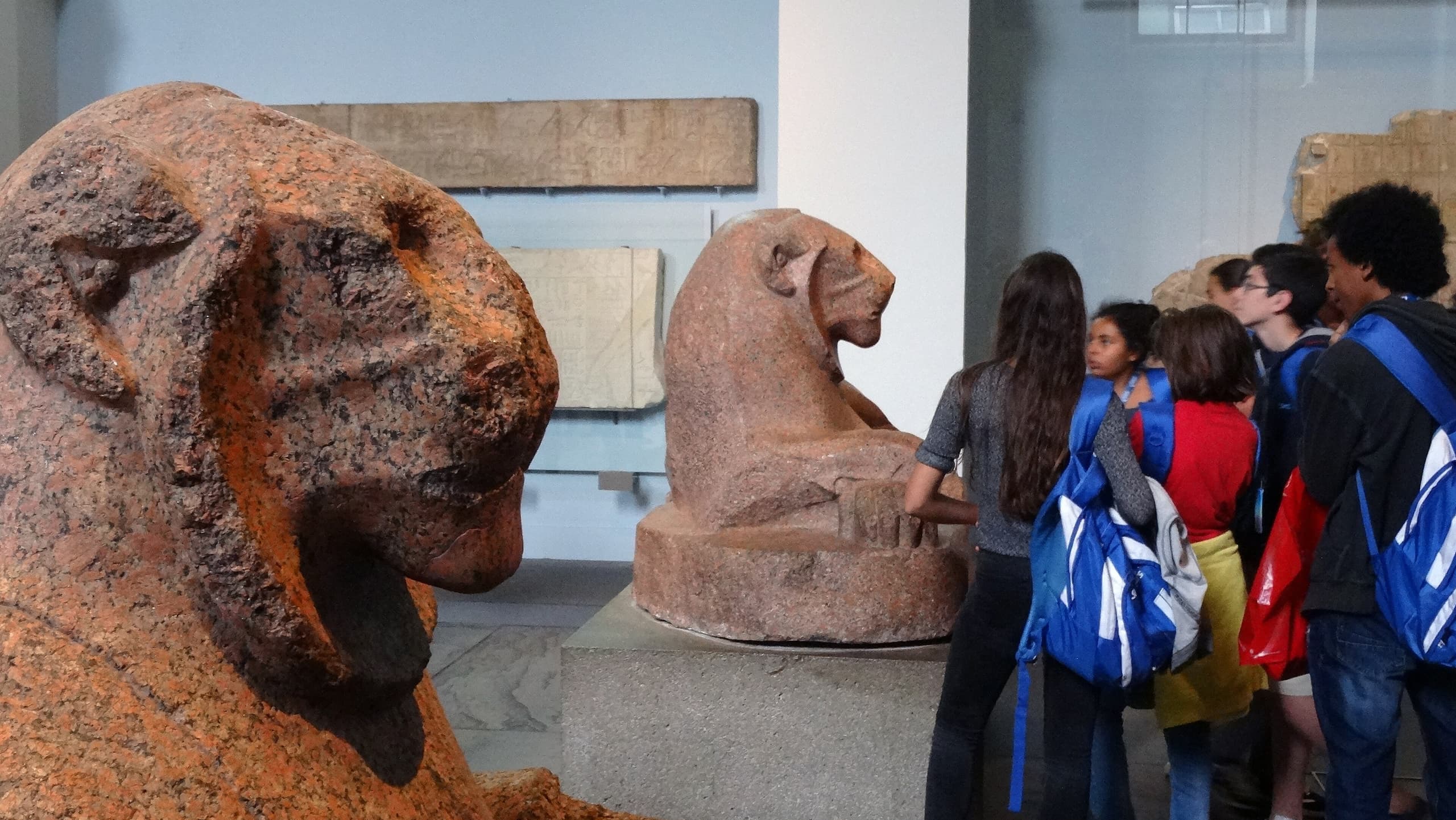A large red granite lion sculpture lying on its side with its head turned and front paws crossed, the surface bearing carved hieroglyphic inscriptions.