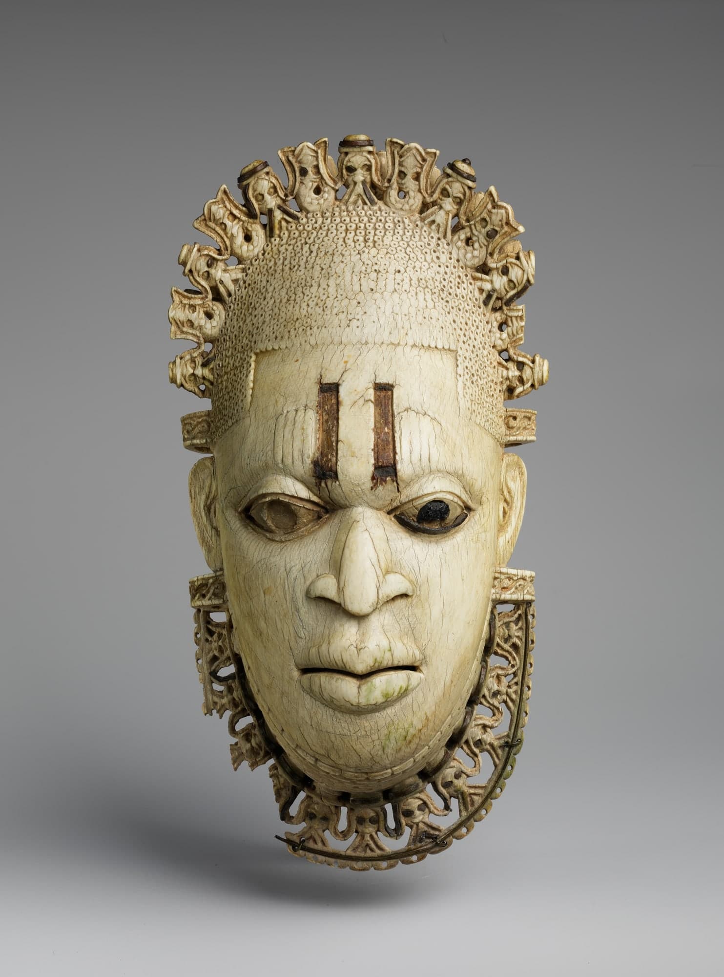 A miniature ivory mask depicting a woman's face with a serene expression, wearing a beaded headdress and choker. An openwork crown rises above, carved with small figures including bearded heads and stylised fish. Three vertical lines mark the forehead.