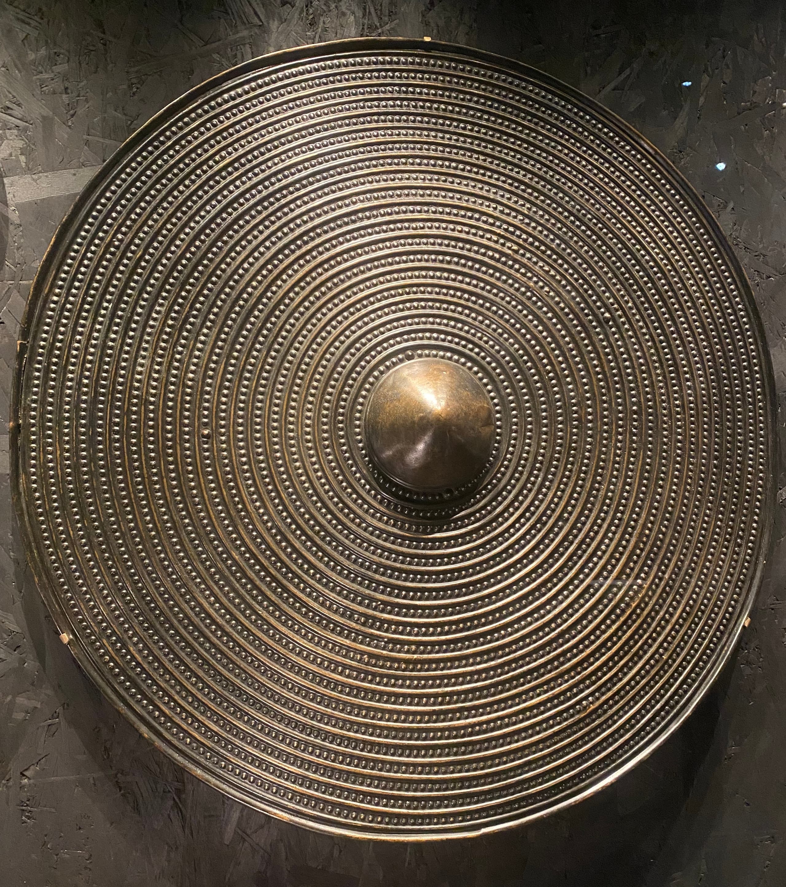A large circular disc of greenish copper alloy with a flat surface and concentric raised ridges radiating from the centre, showing patches of corrosion and patina.