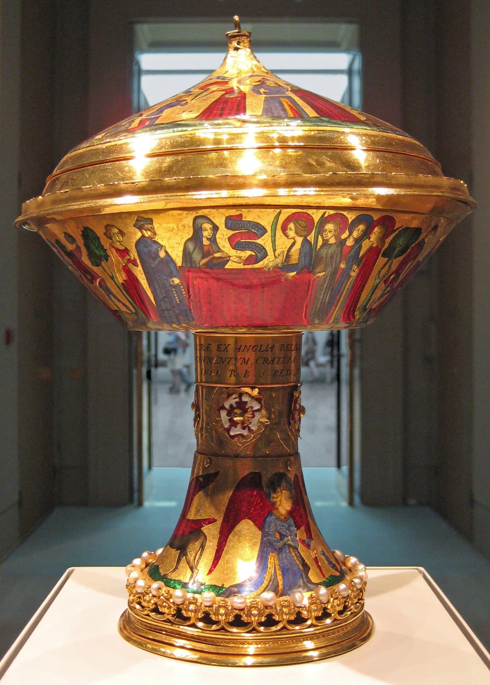 A covered gold cup with a tall stem, decorated with translucent enamel scenes in red, blue and green. Pearls encircle the foot, and the surface is worked with engraved and punched detail.