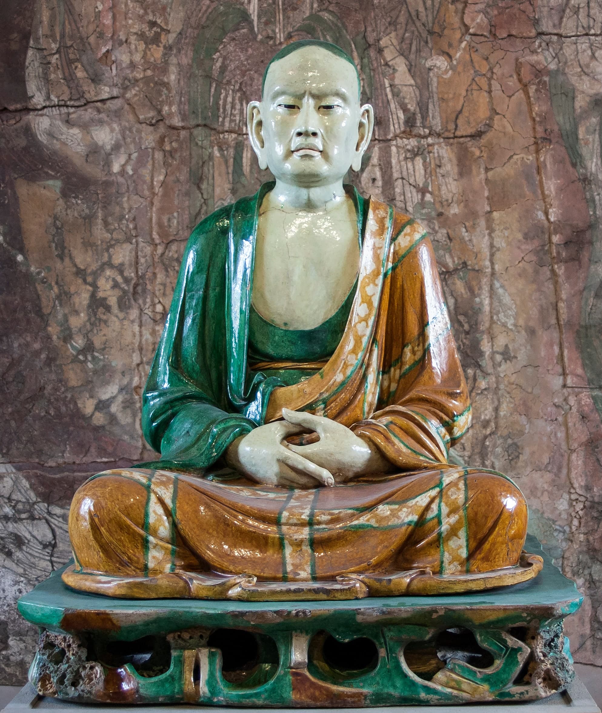 A seated figure in glazed pottery, life-size, with robes draped across the body and hands resting in the lap. The face is naturalistic, with elongated earlobes. The figure sits on an openwork base carved to resemble rock.