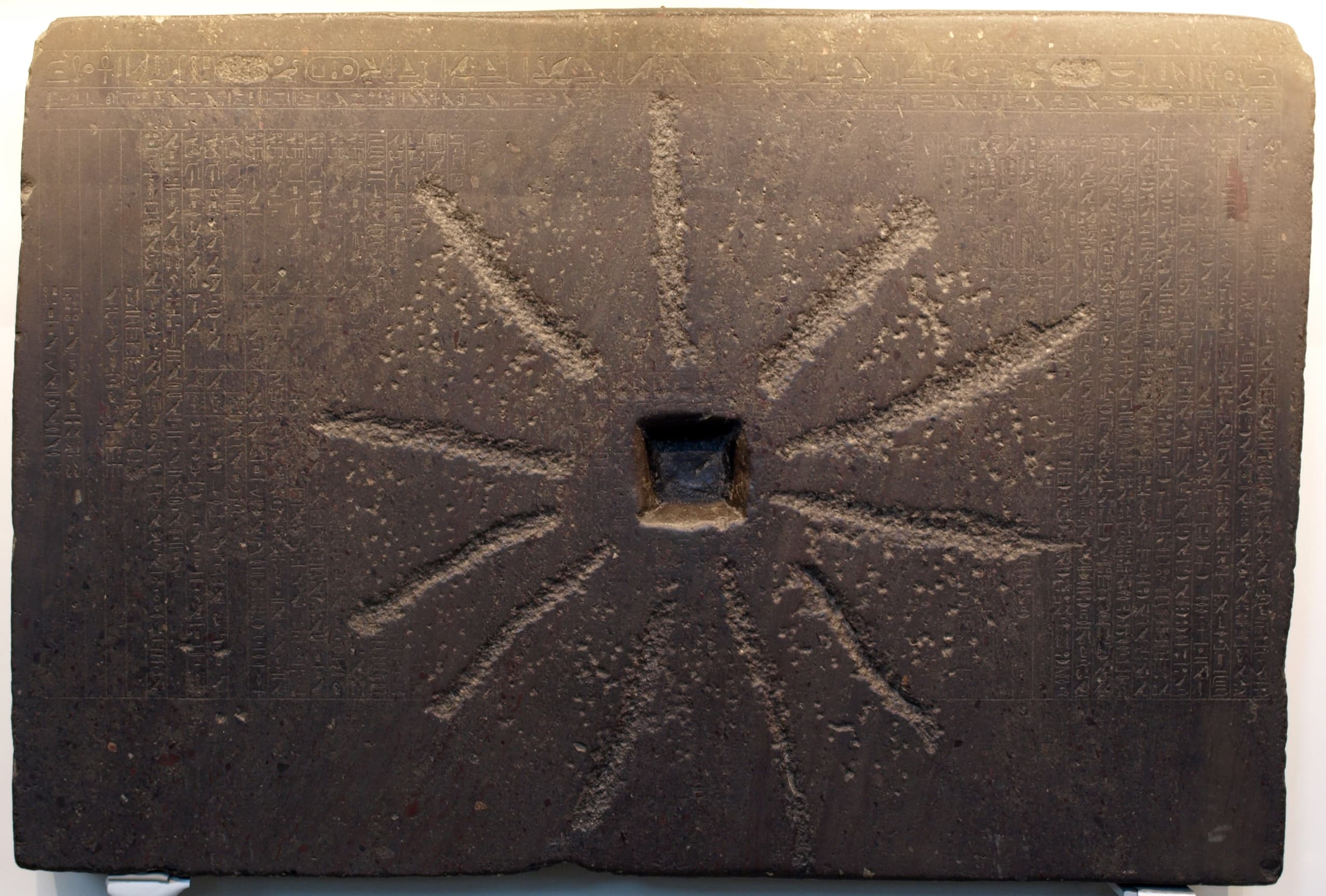 A dark, worn rectangular stone slab whose surface shows broken hieroglyphic text interrupted by heavy abrasion and damaged areas from later reuse.