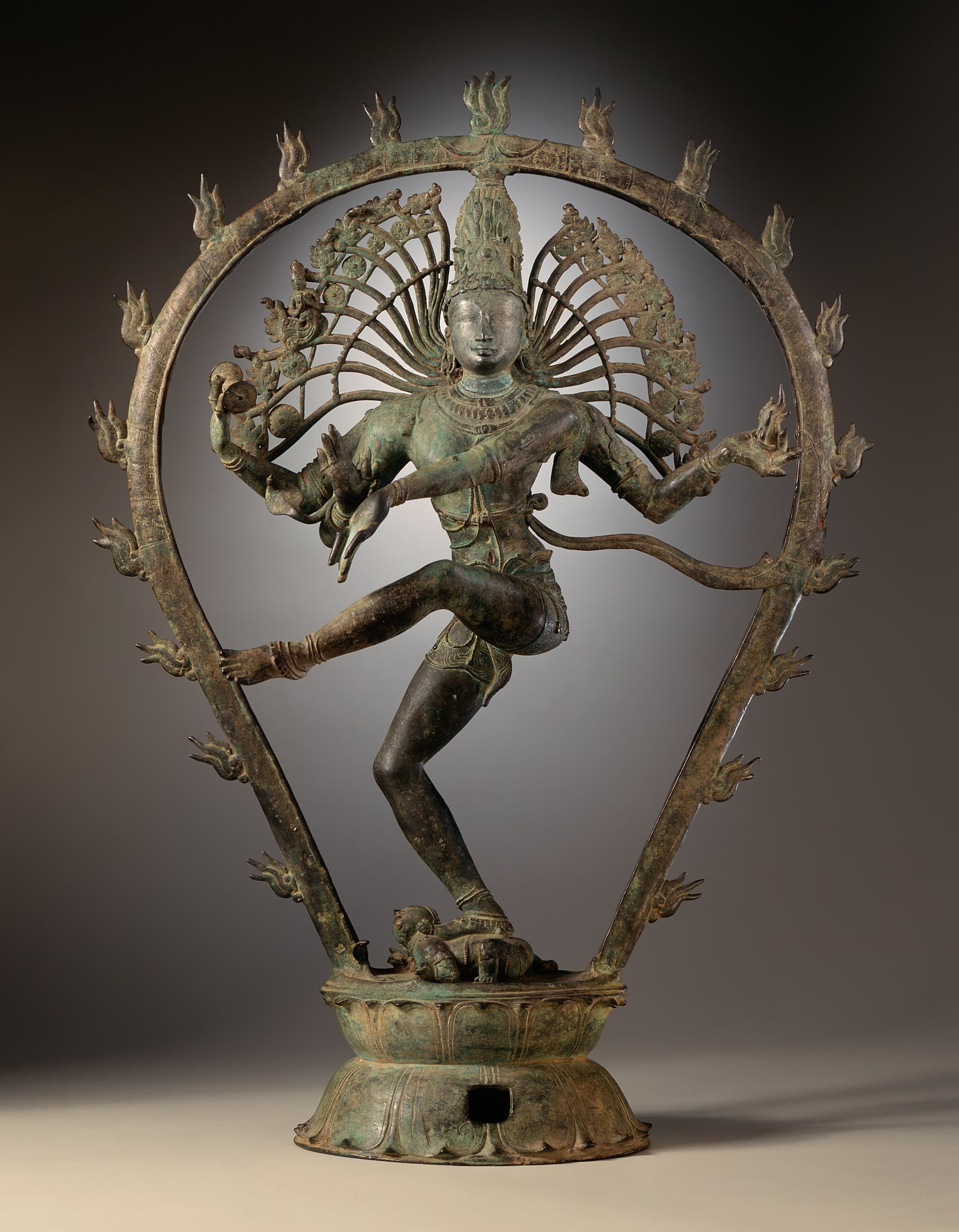 A bronze figure of a four-armed deity dancing on one leg within a ring of flames, trampling a small dwarf figure beneath the raised foot.